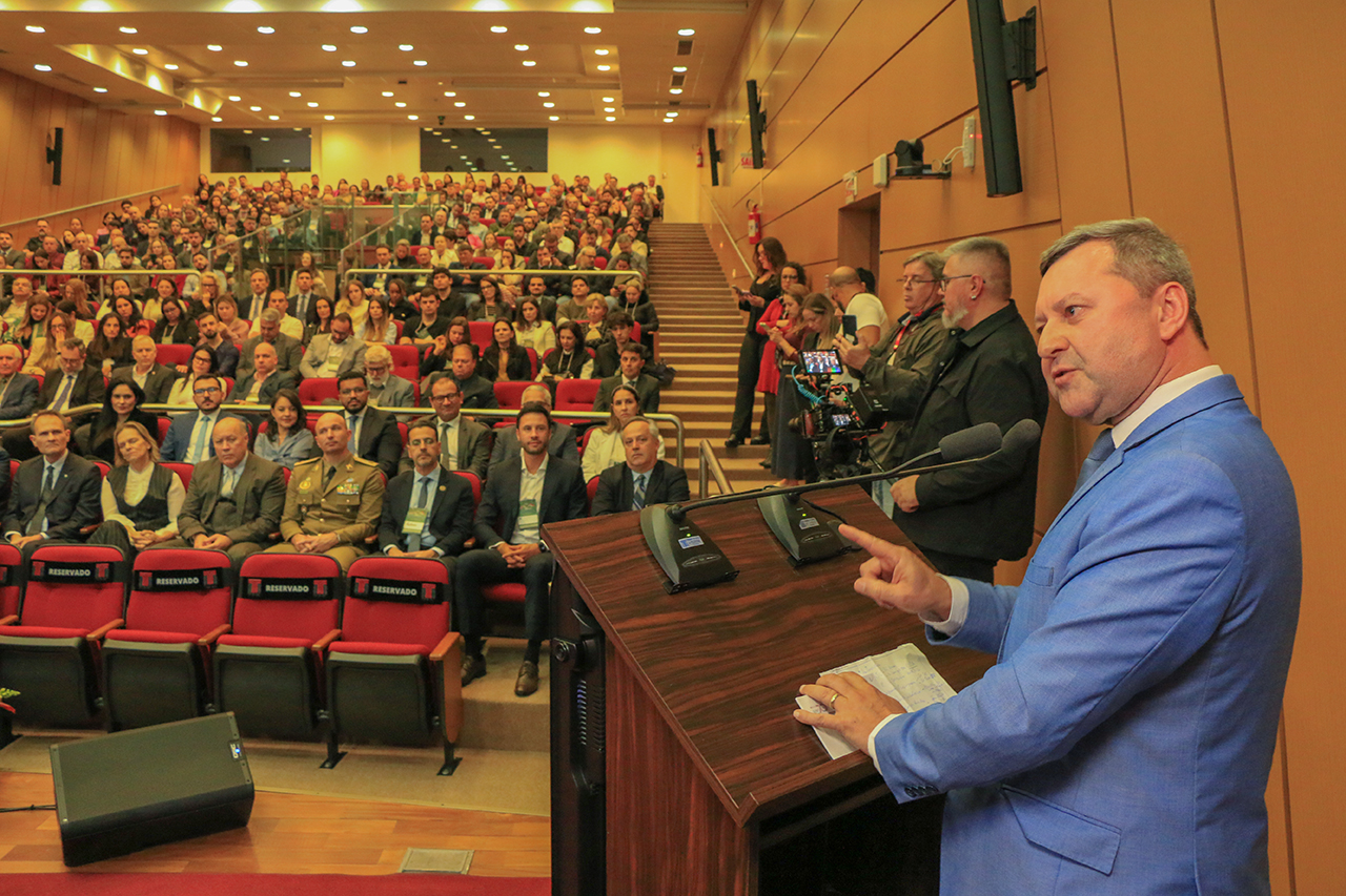 Foto de perfil do presidente do Idasc, José Sérgio Cristóvam, discursando no púlpito, no palco do auditório bordô. Ele é um homem branco, de cabelos grisalhos. Veste terno azul-claro. À sua frente, a plateia que o assiste.