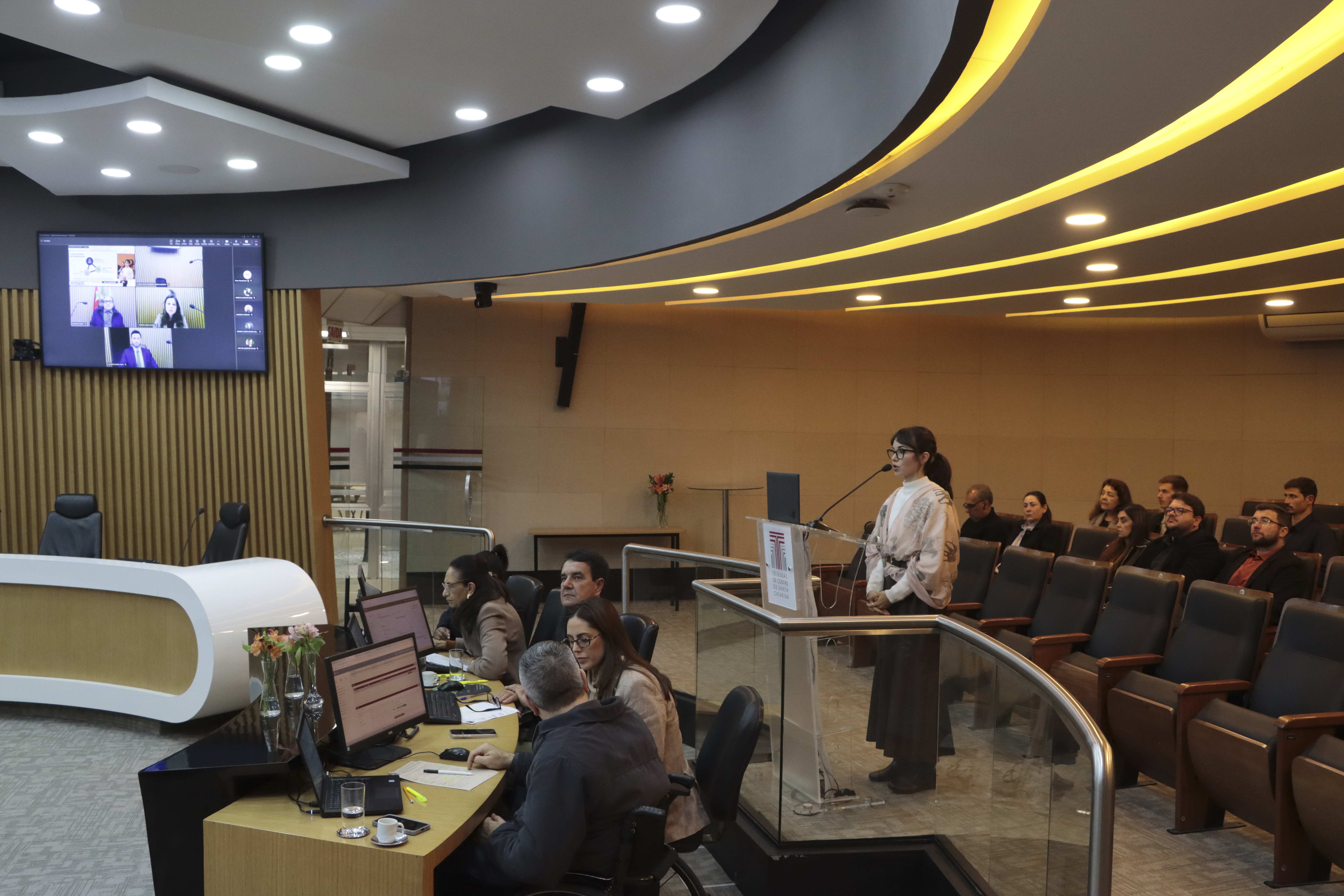 Foto horizontal que mostra a servidora Sabrina Pecini da Silva falando ao púlpito, no Plenário do TCE/SC. É uma mulher branca, de cabelos castanhos e presos. À sua frente, a bancada circular das autoridades, e, atrás, a plateia. 