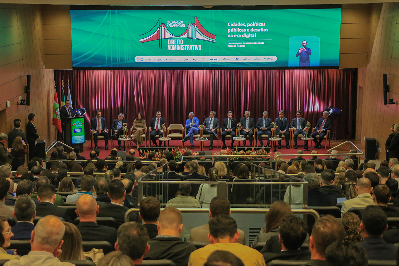 Foto horizontal ampla que mostra o auditório bordô lotado. Aparecem os participantes, de costas, sentados em poltronas enfileiradas, e, no palco, as autoridades durante a abertura do evento. No púlpito, o conselheiro Adircélio de Moraes Ferreira Júnior.