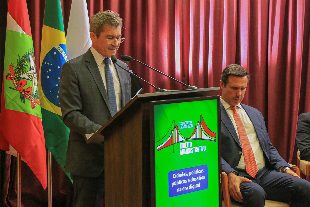 Foto do conselheiro José Nei Ascari discursando no púlpito do auditório. Ele é um homem branco, de cabelos claros. Veste terno e gravata e usa óculos de grau. Na parte da frente do púlpito, é projetado o logo do Congresso. Ao lado, as demais autoridades.