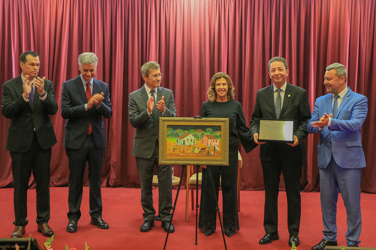 Foto horizontal da cerimônia de homenagem realizada no palco do auditório bordô. O desembargador Ricardo Roesler, do Tribunal de Justiça de Santa Catarina, recebe uma homenagem enquanto segura uma placa comemorativa. Ao centro, um quadro artístico apoiado em um cavalete. Também participam da solenidade os conselheiros José Nei Ascari e Adircélio Ferreira Júnior, o desembargador Rodrigo Collaço, além de outras autoridades, todos em trajes formais.