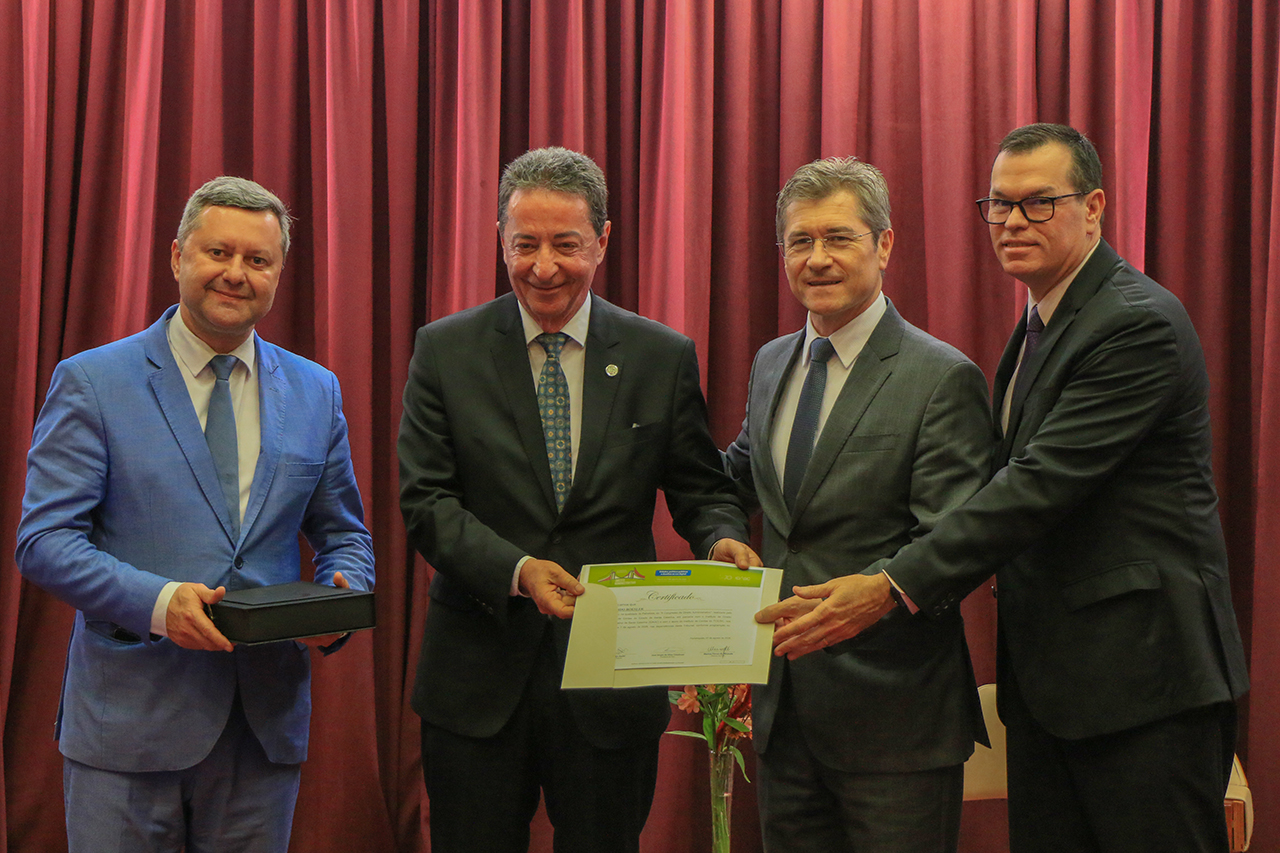 Foto horizontal de autoridades durante a homenagem. Ao centro, o desembargador Ricardo Roesler, do TJSC, recebe uma homenagem e segura, juntamente com os conselheiros José Nei Ascari e Adircélio Ferreira Júnior, uma placa comemorativa. À esquerda, José Sérgio Cristóvam participa da solenidade. Todos estão vestidos em trajes formais e posam para a foto.