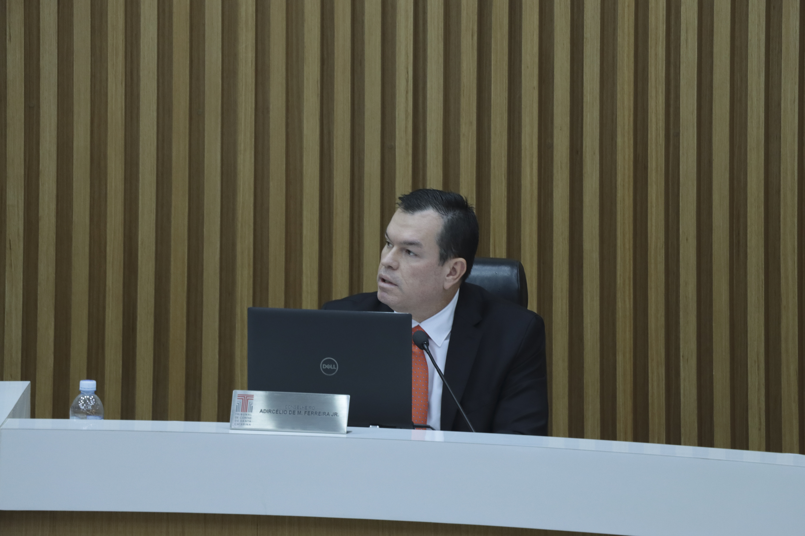 Foto horizontal do plenário do TCE/SC, com o conselheiro Adircélio sentado à bancada, utilizando um notebook e microfone durante a sessão. Ele aparece de perfil, falando sobre a apresentação da servidora Julia, em ambiente formal com painel de madeira ao fundo. 