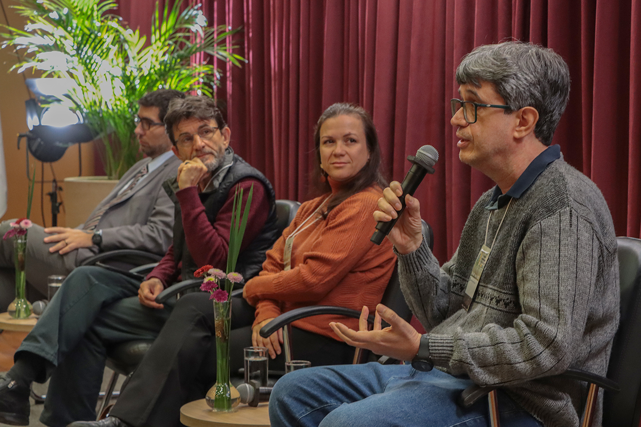 Foto horizontal de quatro painelistas no palco do auditório do TCE/SC, sentados, lado a lado, em poltronas. Fala ao microfone o engenheiro agrônomo José Espíndola. Ao seu lado, a agente do PNAE Manuella Machado e o economista Gustavo Noronha.