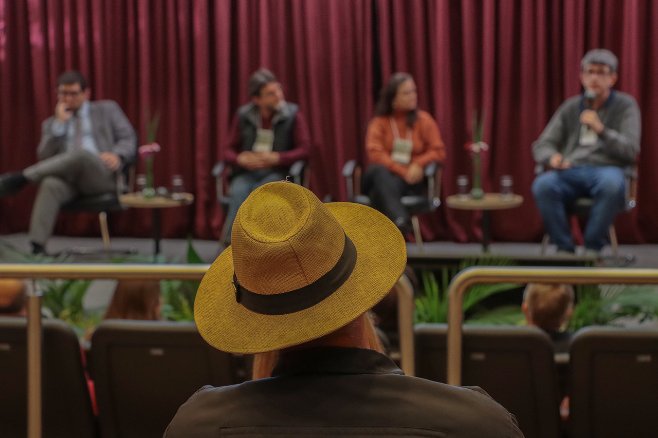 Foto que mostra em primeiro plano uma pessoa de costas, na plateia, que usa um chapéu amarelo. Em plano desfocado, ao fundo, quatro painelistas no palco do auditório do TCE/SC.