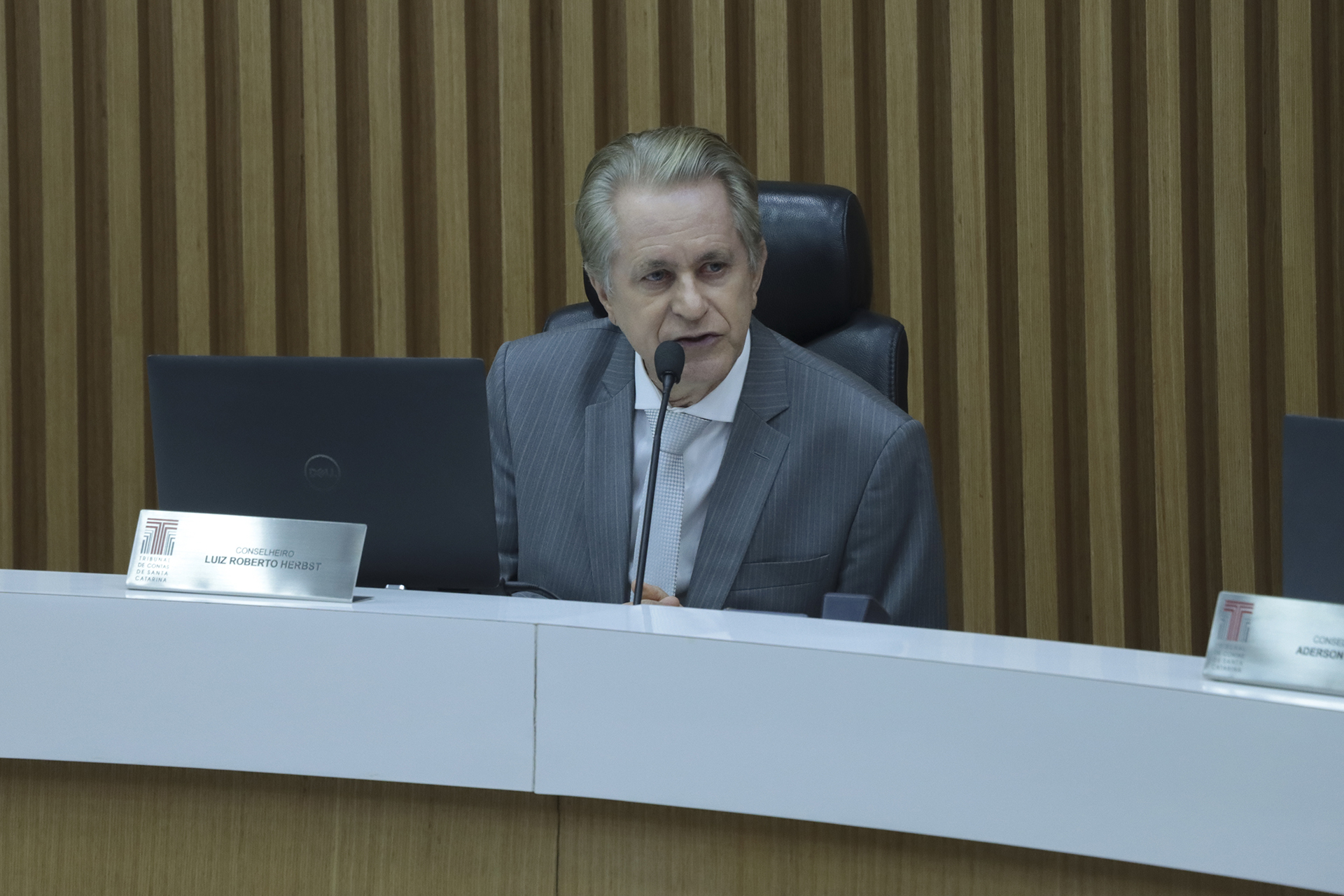 Banner horizontal com foto do conselheiro Luiz Roberto Herbst falando aos demais na sessão plenária. Ele é um homem branco, de cabelos claros. Veste terno e gravata e está sentado diante da bancada de autoridades. 