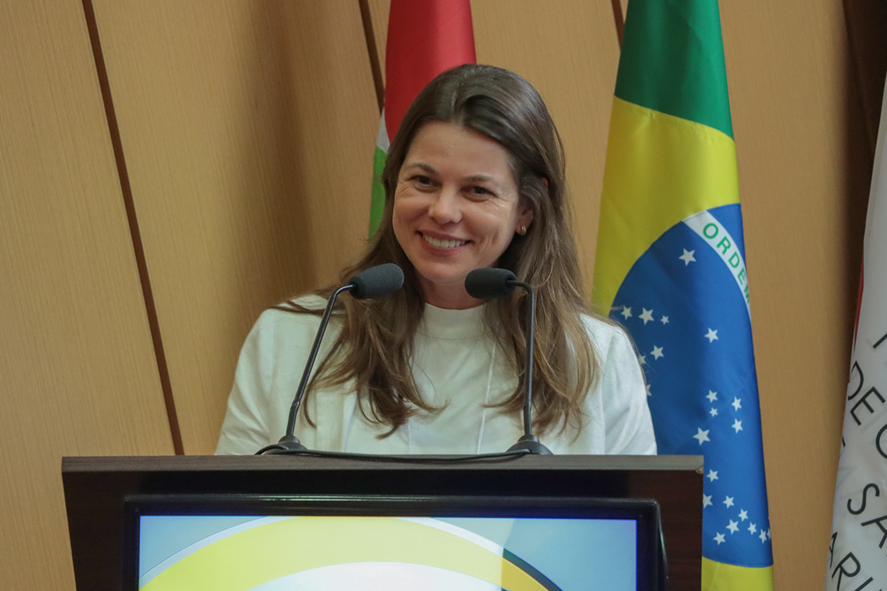 Banner horizontal com foto da conselheira substituta Sabrina Iocken falando ao microfone no púlpito do auditório do TCE/SC. Ela é uma mulher branca, de cabelos castanhos e compridos. Está sorrindo. Ao fundo, painel amadeirado e bandeiras institucionais.
