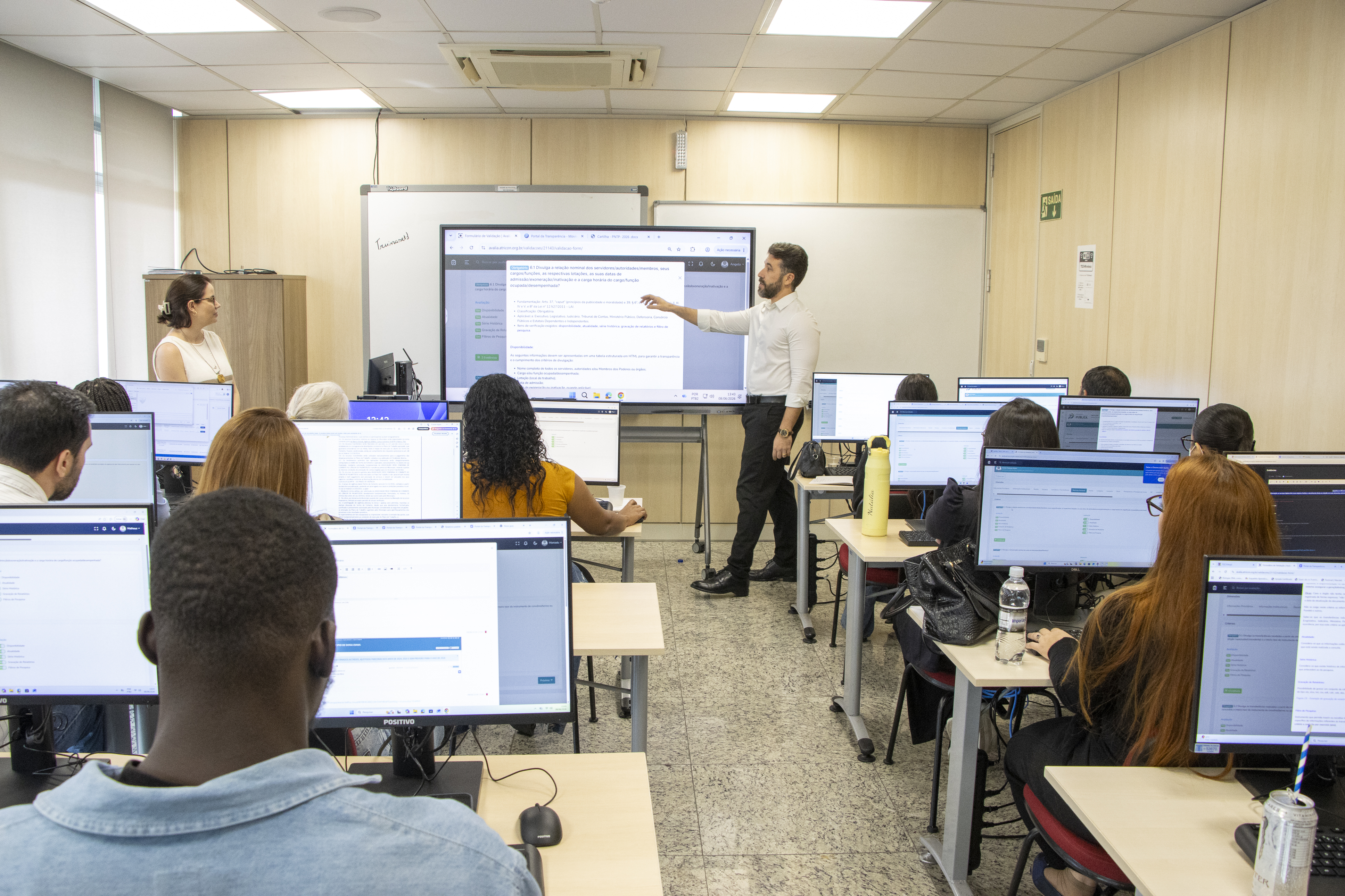 Imagem horizontal de uma capacitação realizada em uma das salas de aula do Instituto de Contas do TCE/SC. Em primeiro plano, participantes, de costas, sentados em frente a computadores, acompanhando o treinamento. Ao fundo da sala, os auditores fiscais de Controle Externo Michelle Padovese de Arruda e Jean Rodrigues de Souza conduzem a atividade. 