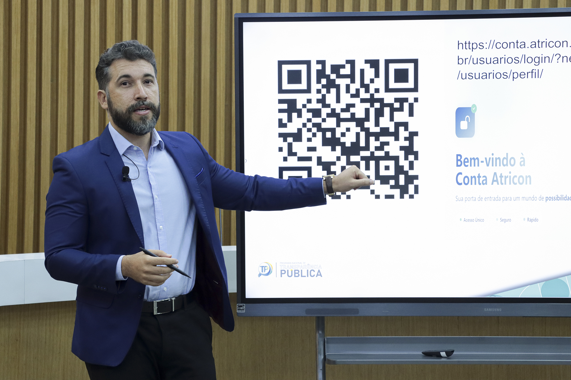 Foto horizontal do auditor fiscal de Controle Externo Jean Rodrigues de Souza no Plenário do TCE/SC, vestindo terno azul e camisa clara, apresentando ao lado de um telão que exibe um QR code. Ele gesticula em direção à tela enquanto conduz a explicação, em cenário com painel de madeira ao fundo.