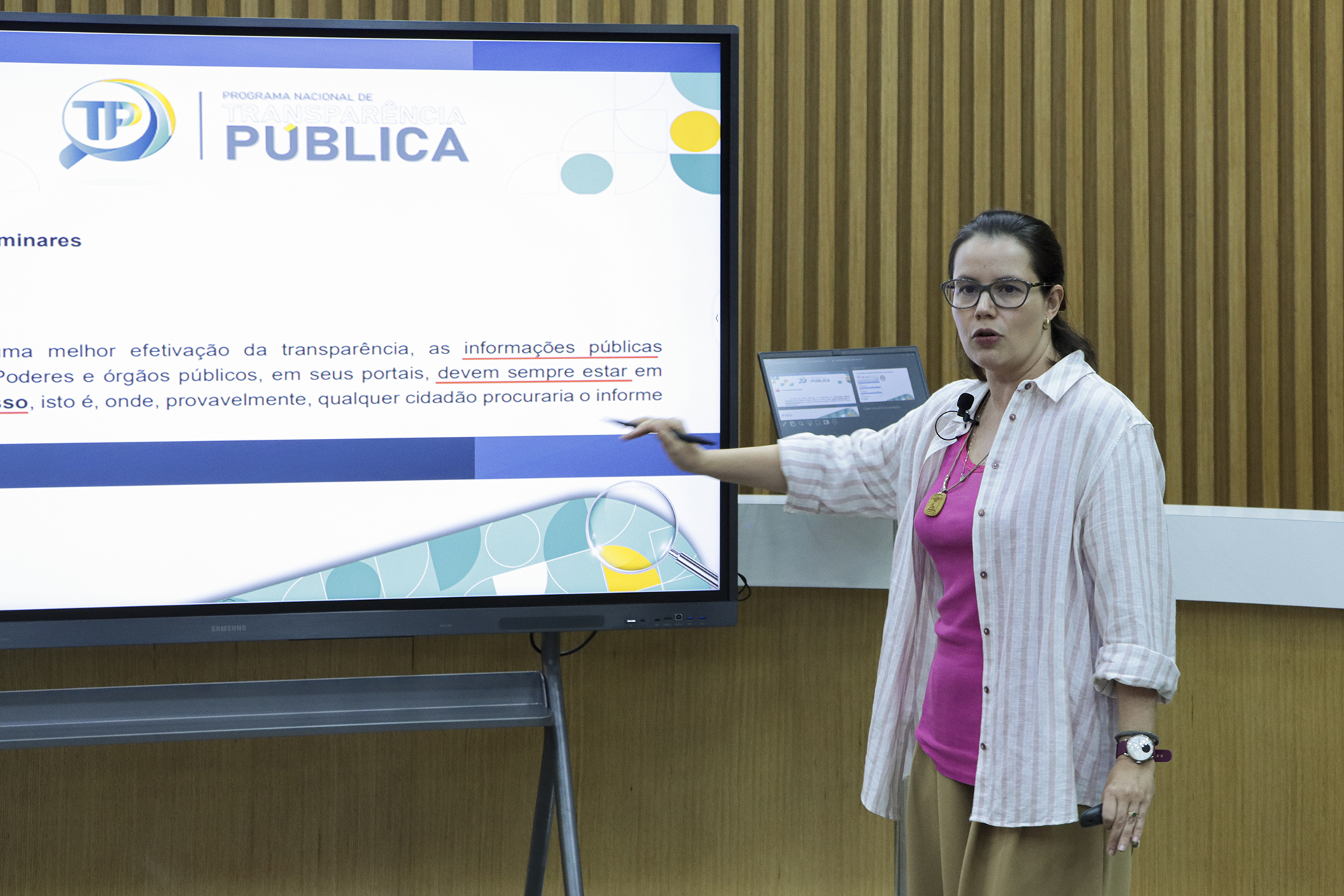 Foto horizontal da auditora fiscal de Controle Externo Michelle Padovese de Arruda durante apresentação no PNTP. Ela é uma mulher branca, de cabelos escuros. Está em pé, em frente a um televisor que exibe a apresentação, falando ao microfone. Veste roupa clara. Está em ambiente institucional com painel de madeira ao fundo.