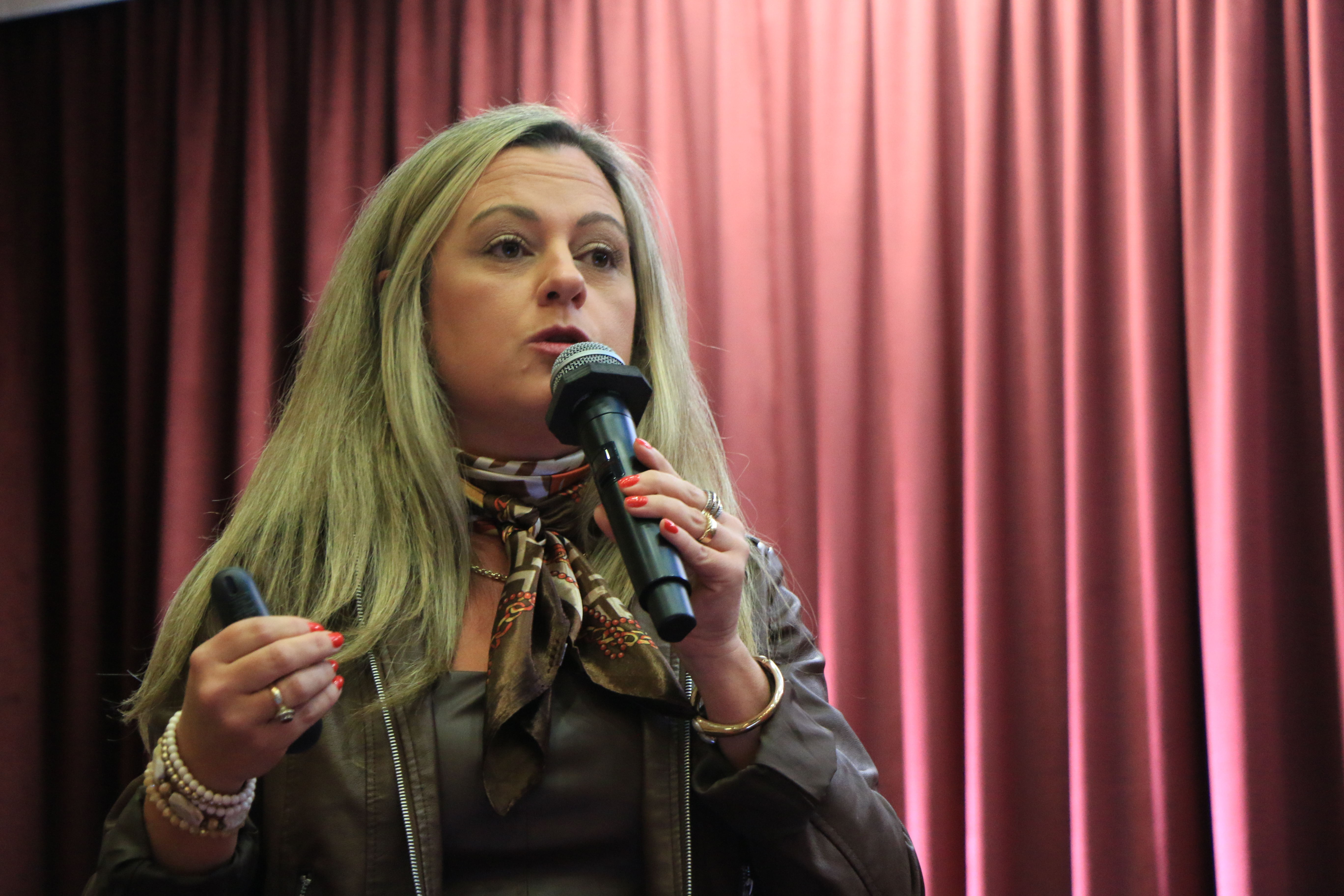 Foto da advogada Caroline Bitencourt falando ao microfone durante o evento. É uma mulher branca, de cabelos claros. Está em pé e veste roupa escura. Ao fundo, cortinas bordôs. 