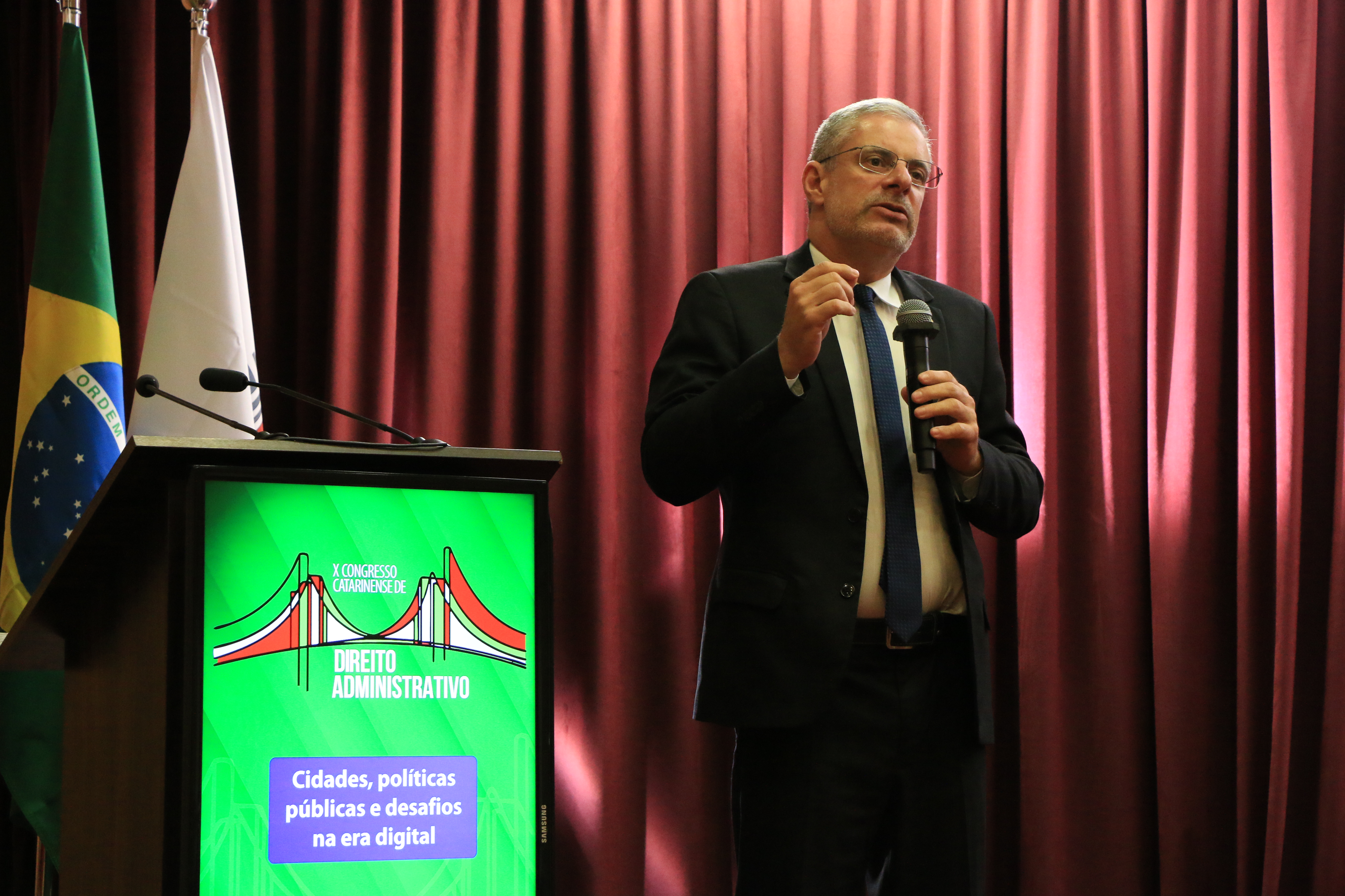 Foto horizontal do conselheiro substituto Gerson Sicca falando ao microfone durante o evento. É um homem branco, de cabelos grisalhos. Veste terno e gravata e usa óculos de grau. Ele está próximo ao púlpito, que exibe o logo do congresso. 