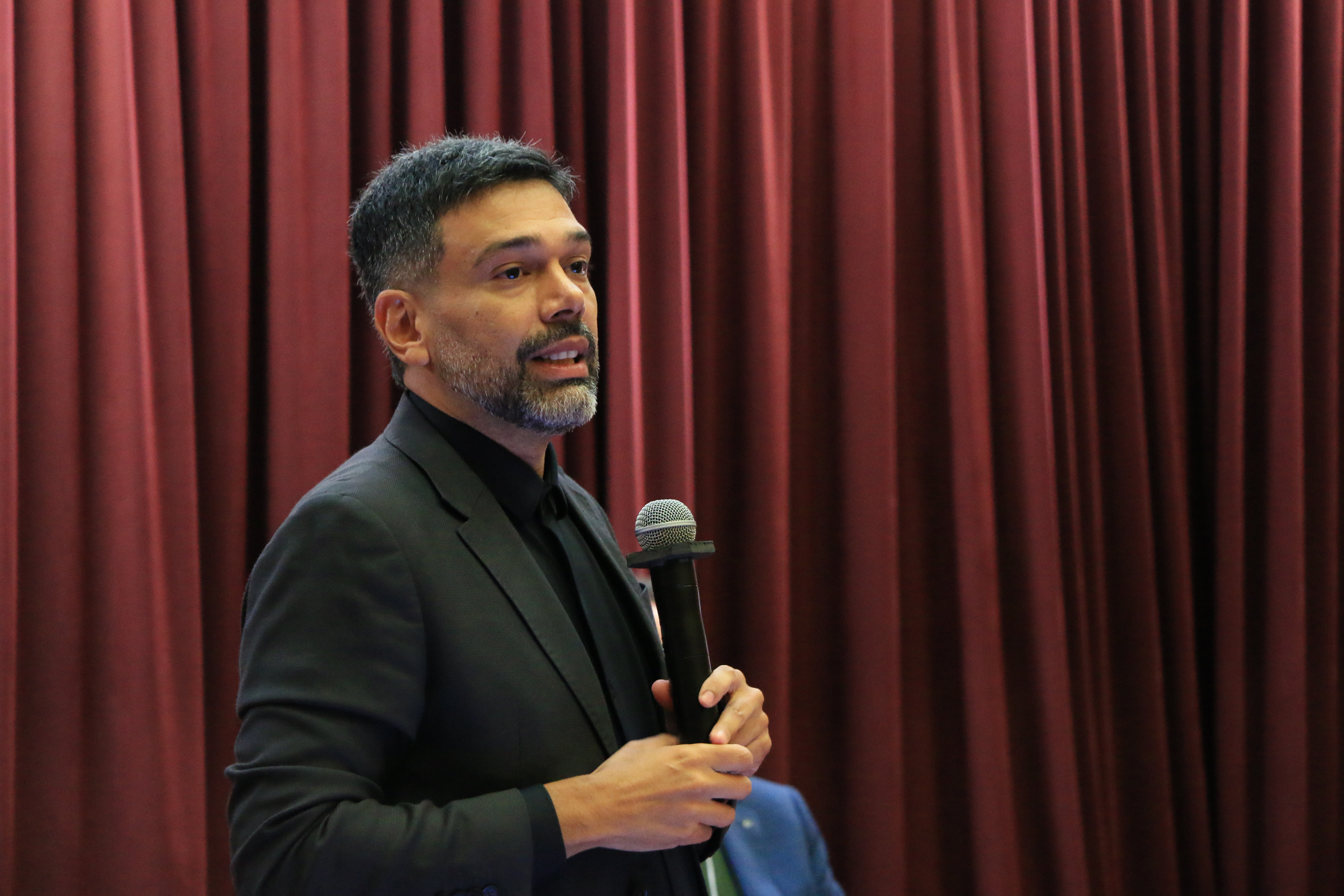 Foto horizontal do auditor Fabian Maia falando ao microfone durante o evento. É um homem branco, de cabelos e barba escuros. Veste paletó preto e está em pé. O cenário ao fundo é composto por cortinas bordôs.