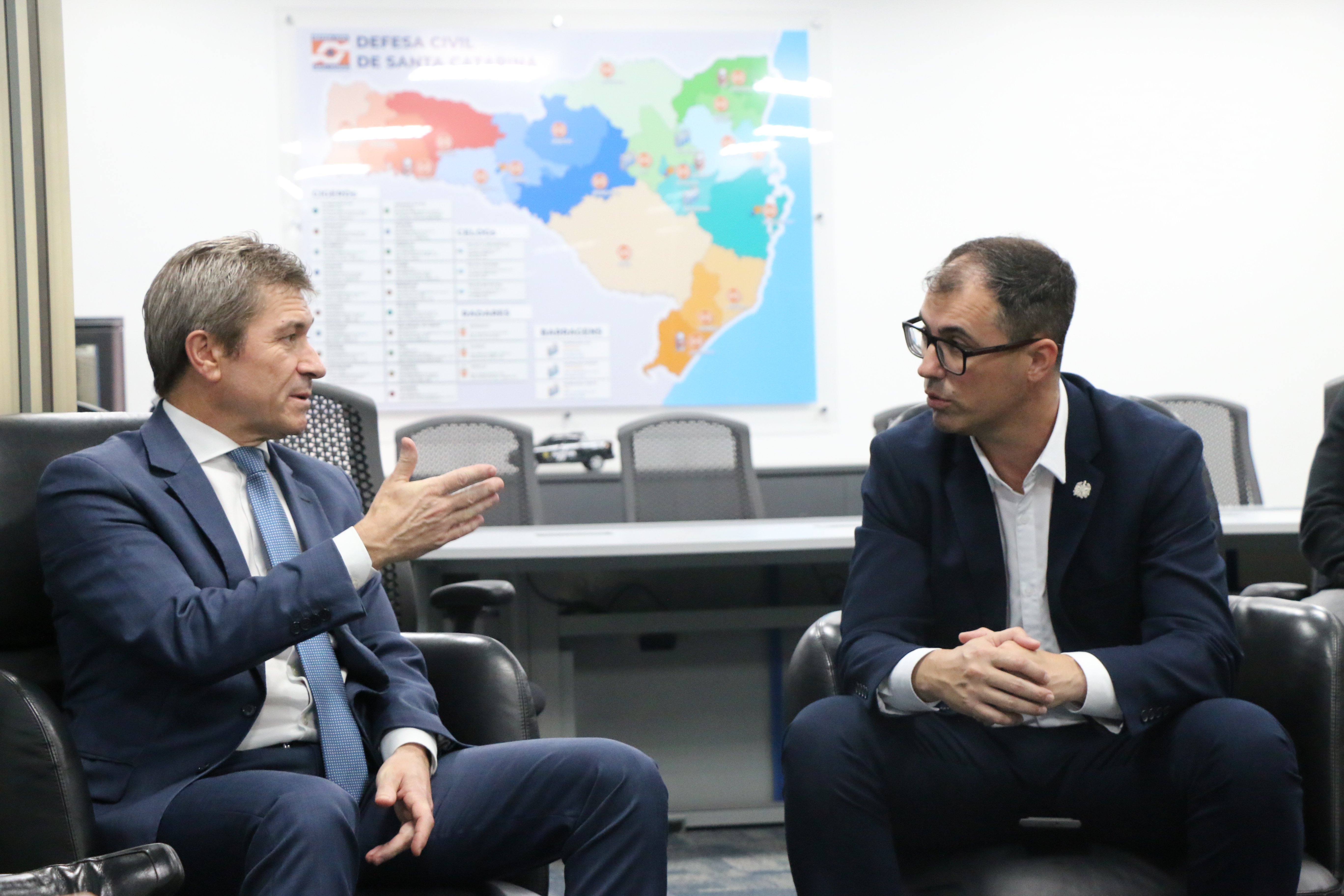 Banner horizontal com foto do conselheiro José Nei Ascari (à esquerda) e do secretário da Proteção e Defesa Civil do Estado, Fabiano de Souza (à direita). Ambos estão sentados em poltronas, um ao lado do outro, enquanto conversam. Vestem terno e gravata. Ao fundo, na parede, há um desenho do mapa de Santa Catarina 