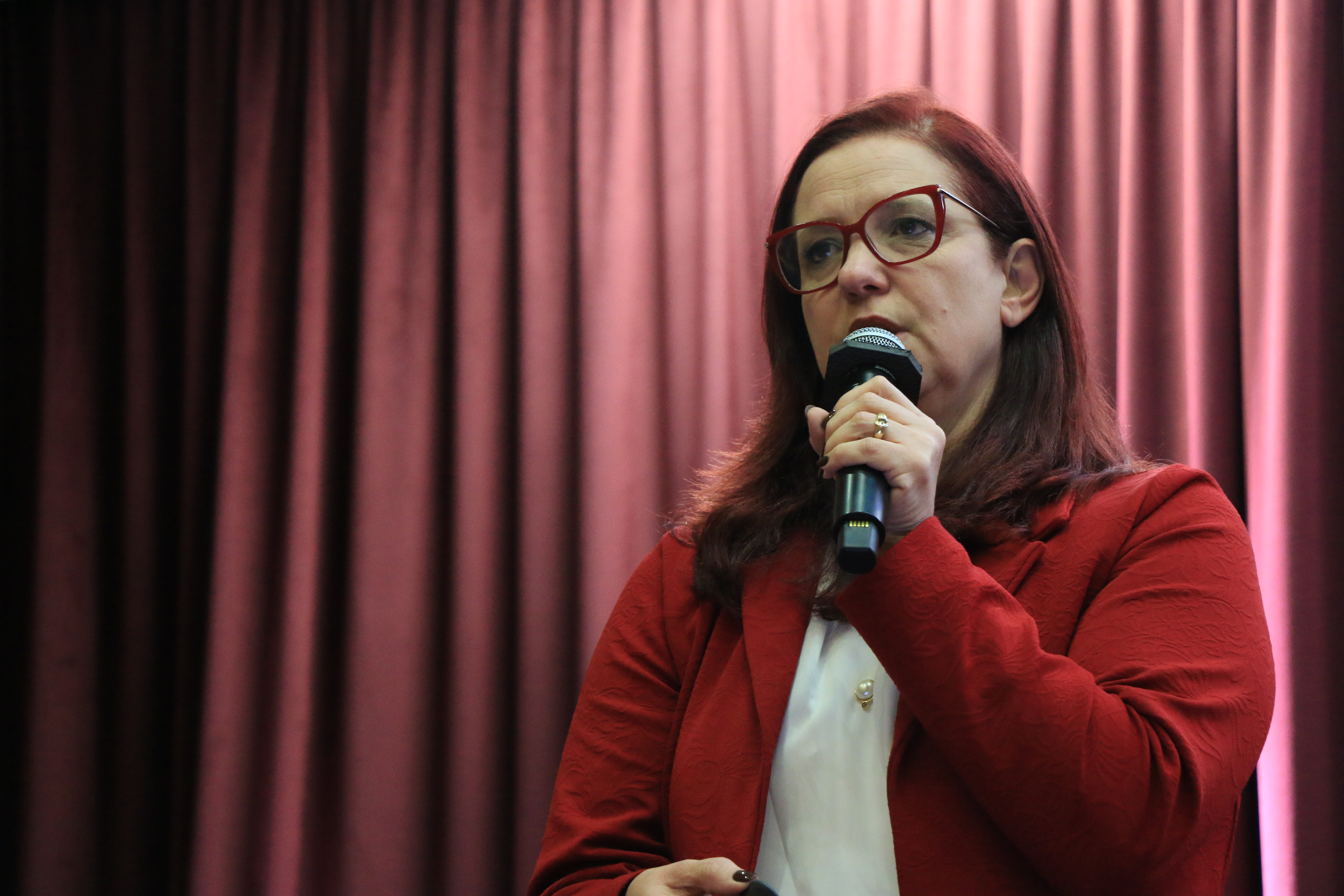 Foto horizontal da presidente do IPDA, Adriana Schier, falando ao microfone durante o evento. É uma mulher branca, de cabelos vermelhos. Veste roupa vermelha. Ao fundo, cortinas bordôs. 