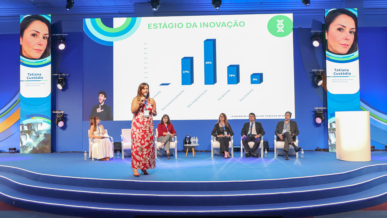 Foto da diretora do LINCE, Tatiana Custódio. Ela está ao lado direito da foto, sobre um tablado. À sua frente, uma plateia formada homens e mulheres a escuta.