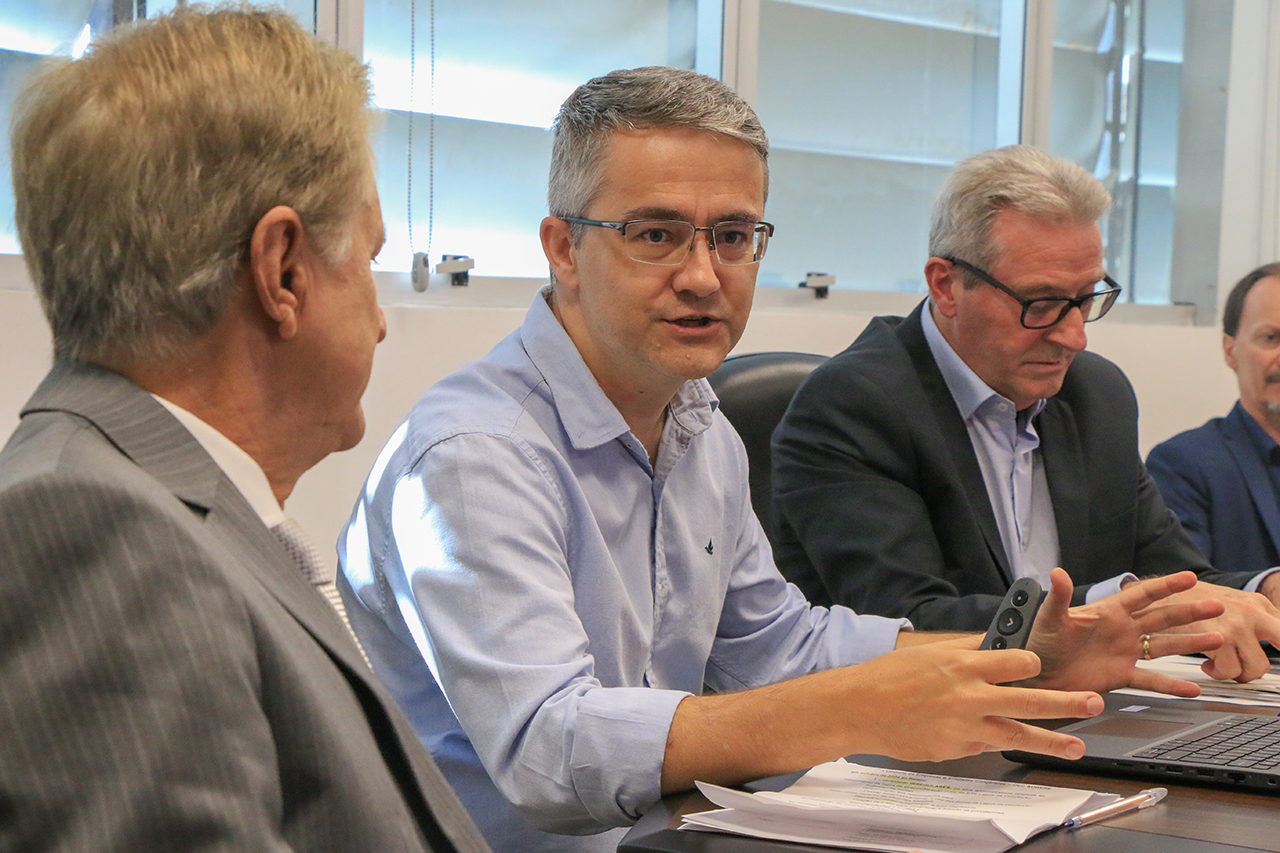 Foto do diretor da DEC, Maximiliano Mazera, falando aos demais durante a reunião. Ele é um homem branco, de cabelos grisalhos. Está ao lado do conselheiro Luiz Roberto Herbst e do coordenador da mesa, Neimar Paludo. 