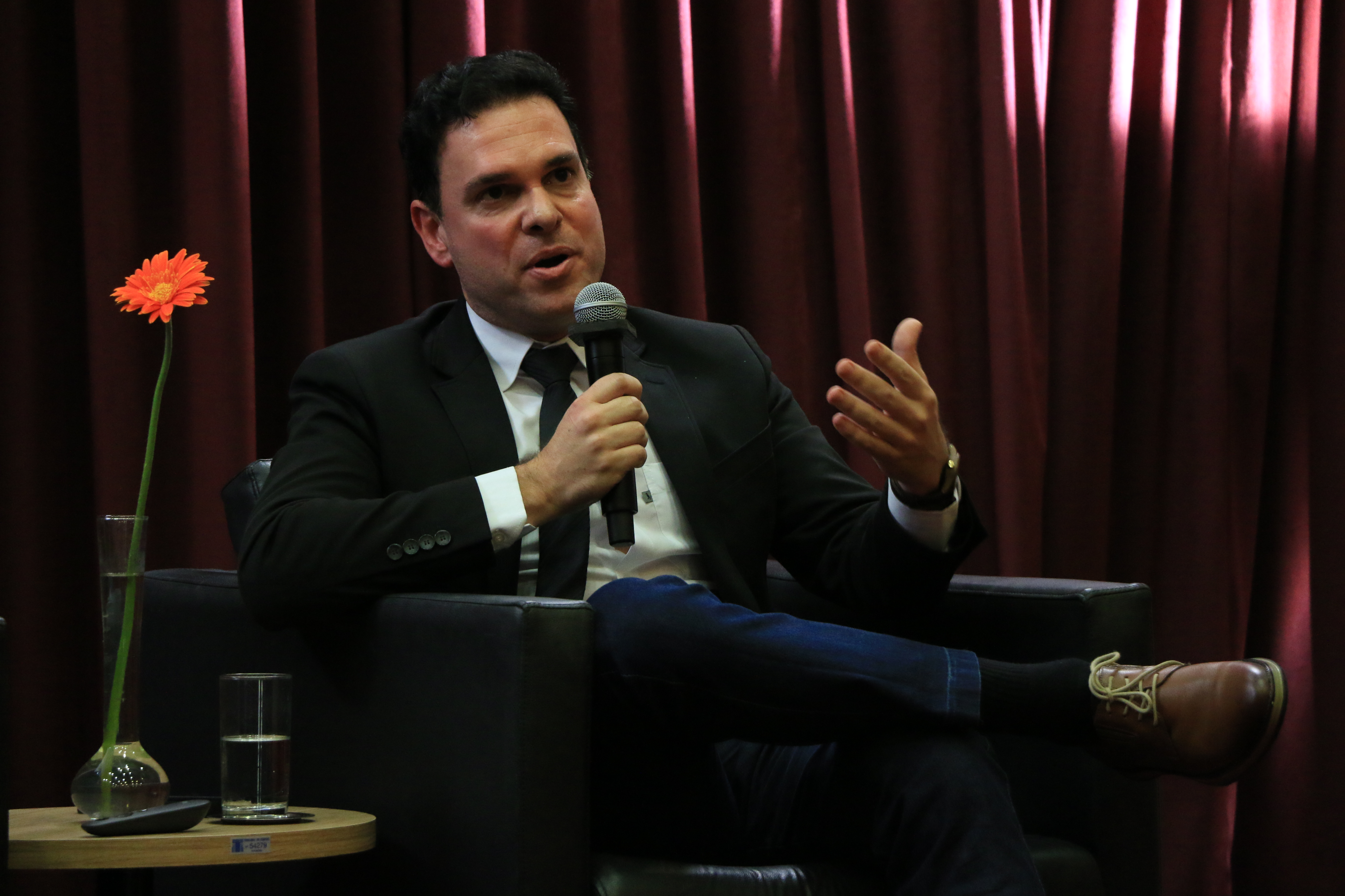 Foto horizontal do procurador Janriê Reck falando ao microfone durante o evento. Ele é um homem branco, de cabelos castanhos. Veste terno e gravata. Está sentado em poltrona no palco. 