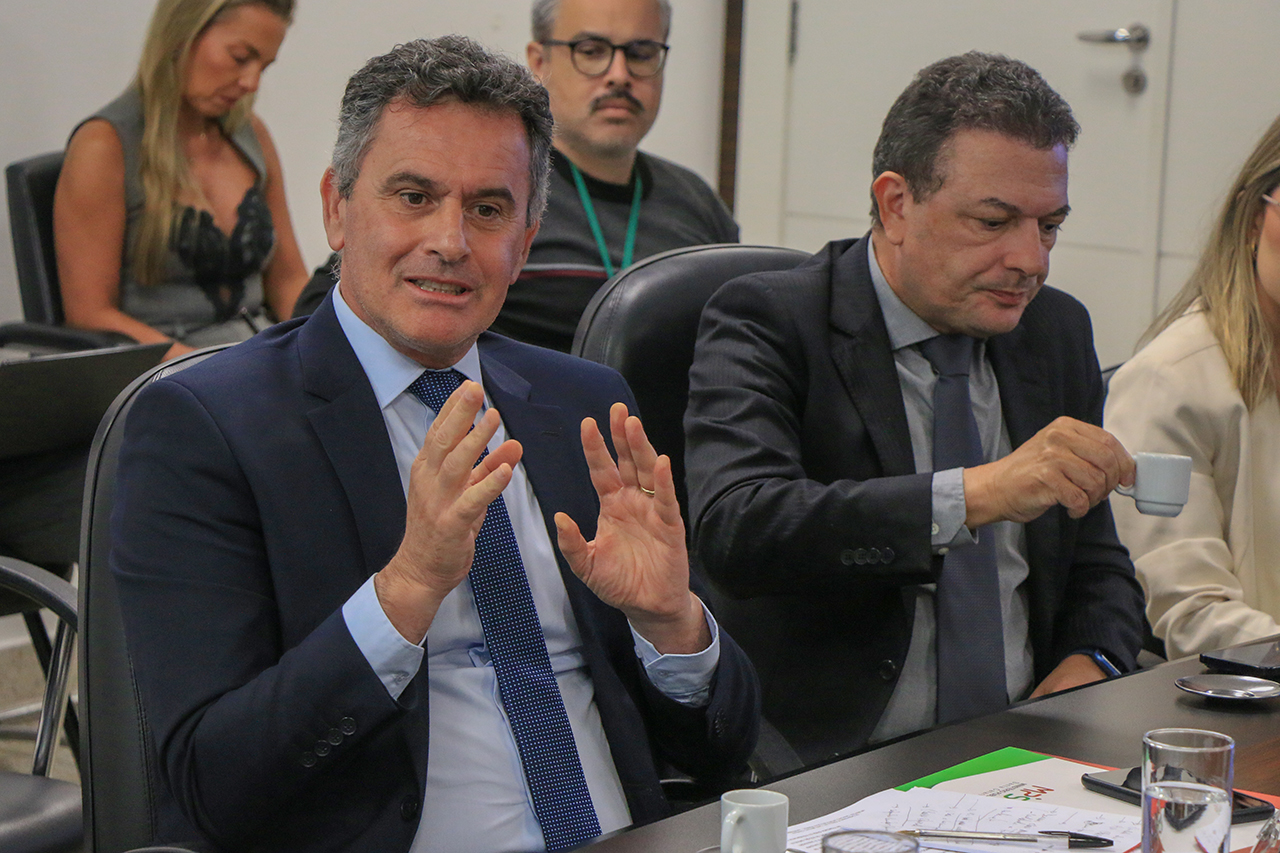 Foto do representante do MPSC, Luiz Fernando Ulysséa, falando aos demais durante reunião. Ele é um homem branco, de cabelos grisalhos. Veste terno e gravata e está gesticulando. Ao seu lado, os outros participantes. 