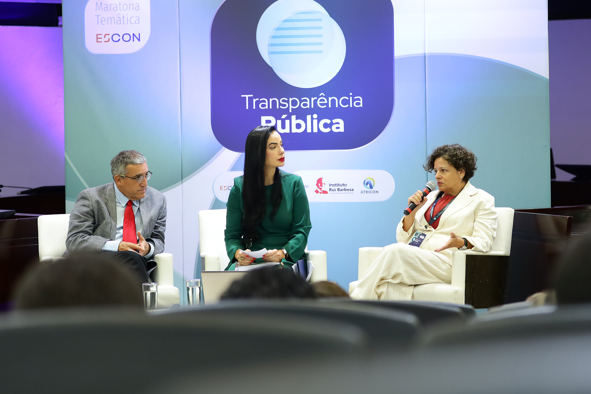Foto horizontal que mostra a auditora fiscal de Controle Externo Joseane Corrêa durante participação em painel, utilizando microfone enquanto se dirige ao público, ao lado de outros participantes sentados em poltronas, em ambiente institucional com painel ao fundo com o tema “Transparência Pública”. 