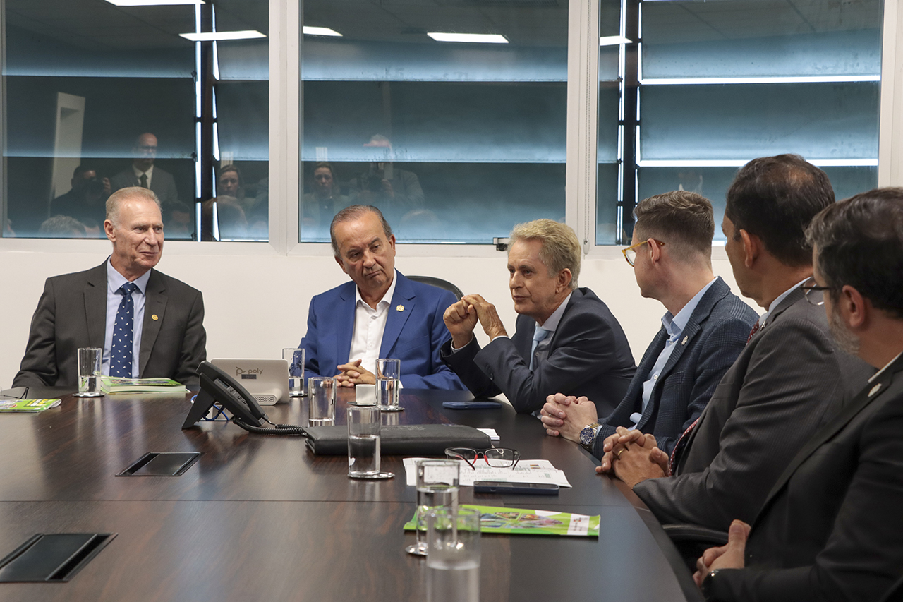 Foto horizontal em ambiente institucional mostrando reunião no TCE/SC, com autoridades sentadas ao redor de uma mesa. Em destaque, o conselheiro-relator das contas de 2025, Luiz Roberto Herbst, fala aos demais. Ao lado, o presidente Herneus De Nadal e o governador Jorginho Mello acompanham atentos, junto a outros participantes, com documentos, copos de água e equipamentos sobre a mesa, em sala com janelas ao fundo.
