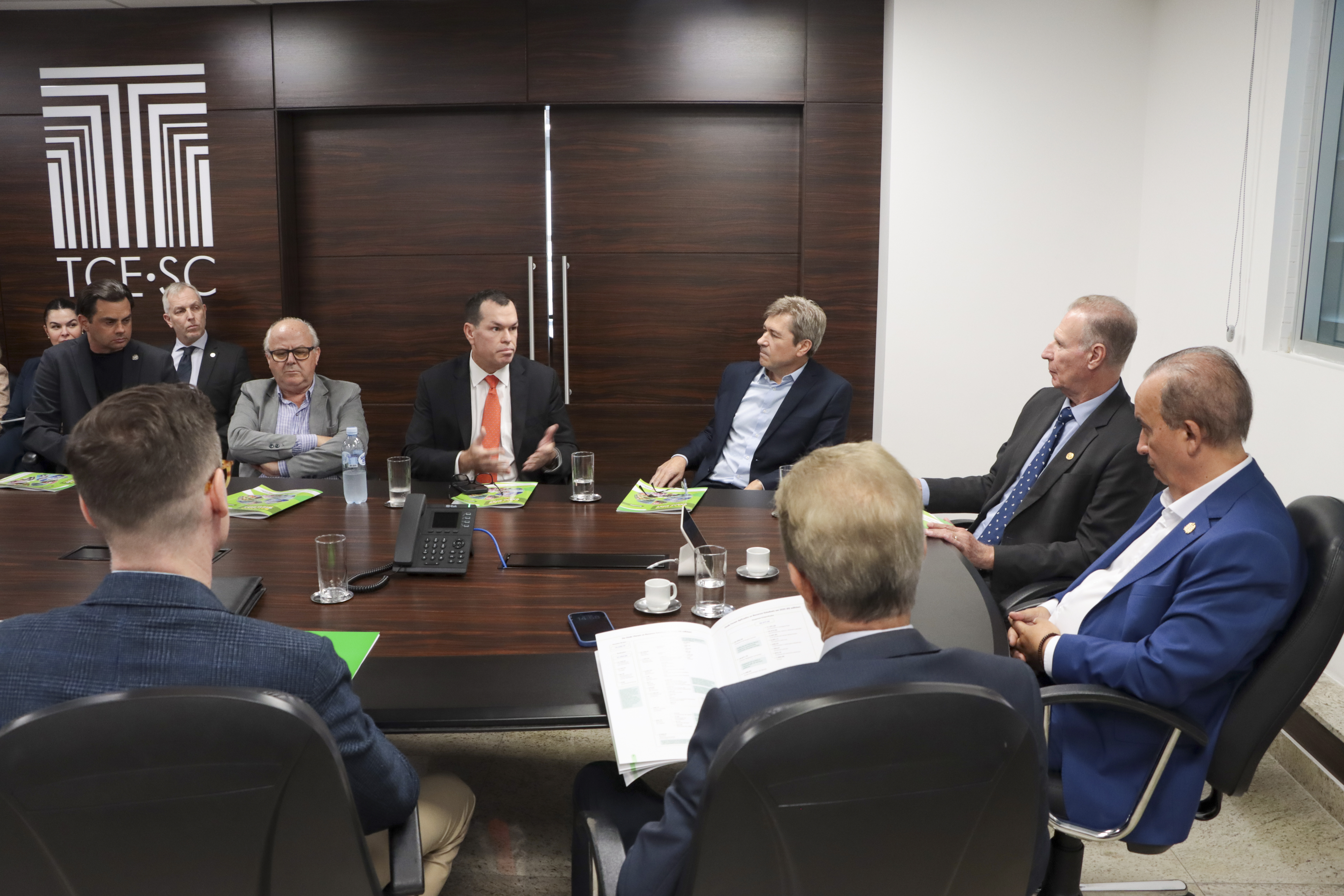 Foto horizontal em ambiente institucional no TCE/SC, mostrando reunião entre autoridades durante a entrega das contas do governo de 2025, com participantes sentados ao redor de uma mesa de madeira escura em uma sala com painel ao fundo exibindo o logotipo do TCE/SC. Entre os presentes, os conselheiros Adircélio de Moraes Ferreira Júnior, José Nei Ascari e Wilson Wan-Dall. Eles estão ao lado dos demais integrantes do TCE/SC, do presidente Herneus De Nadal e do governador Jorginho Mello.