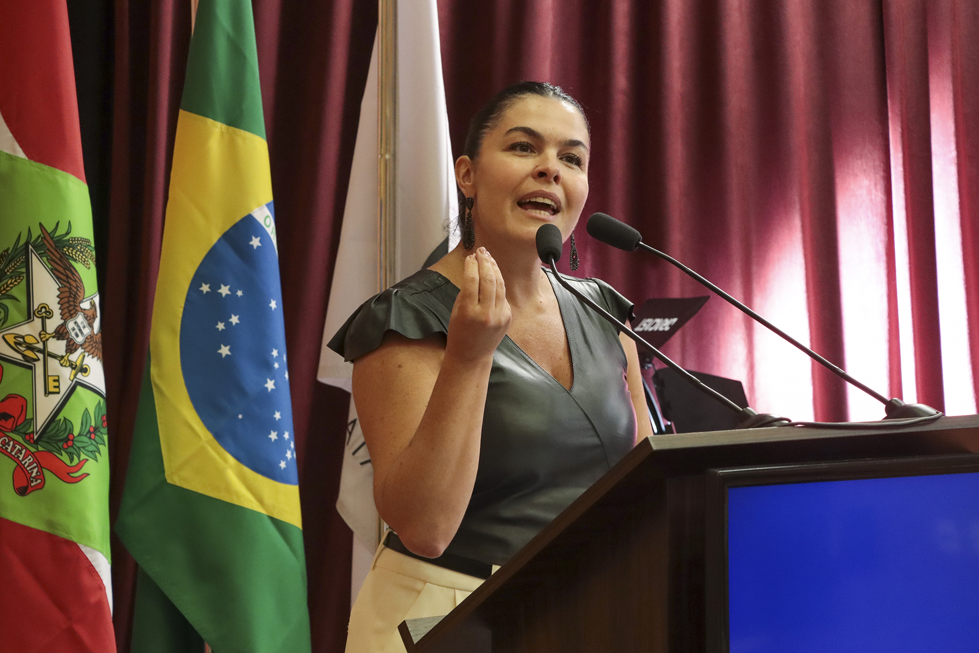 Foto da diretora Monique Portella discursando no púlpito do auditório do TCE/SC durante evento de treinamento, utilizando microfone e gesticulando enquanto se dirige ao público, vestindo traje formal, com bandeiras ao fundo — entre elas a do Brasil e de Santa Catarina — e cortinas vermelhas que compõem o ambiente institucional. Ela é uma mulher branca, de cabelos escuros.