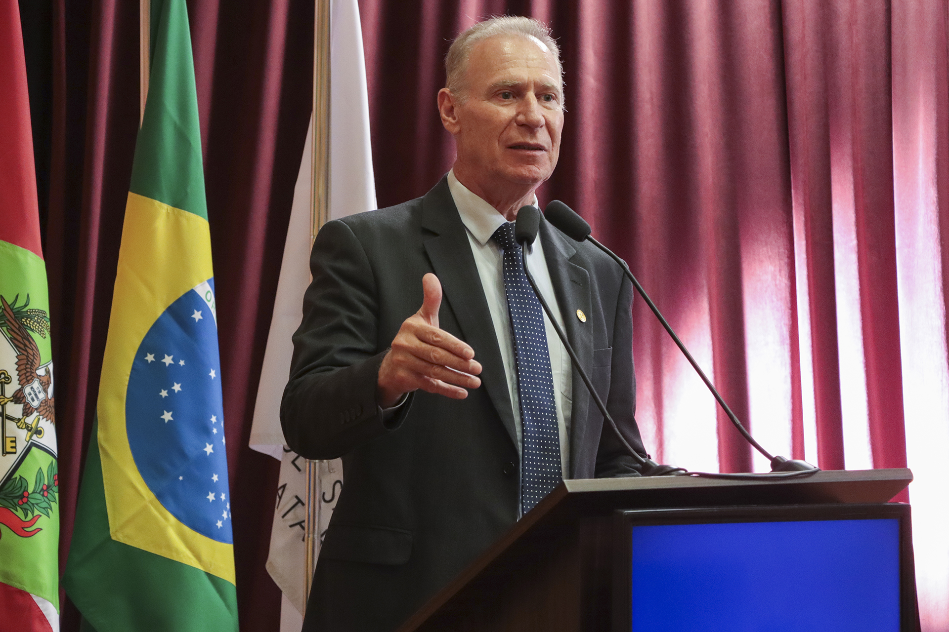 Foto horizontal do presidente do TCE/SC, conselheiro Herneus De Nadal, durante sua fala no púlpito do auditório do TCE/SC, no evento de treinamento, utilizando microfone e gesticulando enquanto se dirige ao público, em ambiente institucional decorado com bandeiras e cortinas vermelhas. Ele é um homem branco, de cabelos claros.