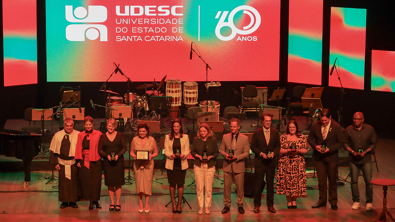 Foto horizontal que mostra 11 pessoas em pé, no palco, uma ao lado da outra. Os presentes na foto são os agraciados com a medalha. Entre eles, a chefe de Gabinete da Presidência do TCE/SC, Juliana Francisconi Cardoso. Estão presentes também o reitor da Udesc, José Fernando Fragalli, e a vice-reitora, Clerilei Aparecida Bier. Ao fundo, no telão, é projetado o banner em comemoração aos 60 anos da Udesc.