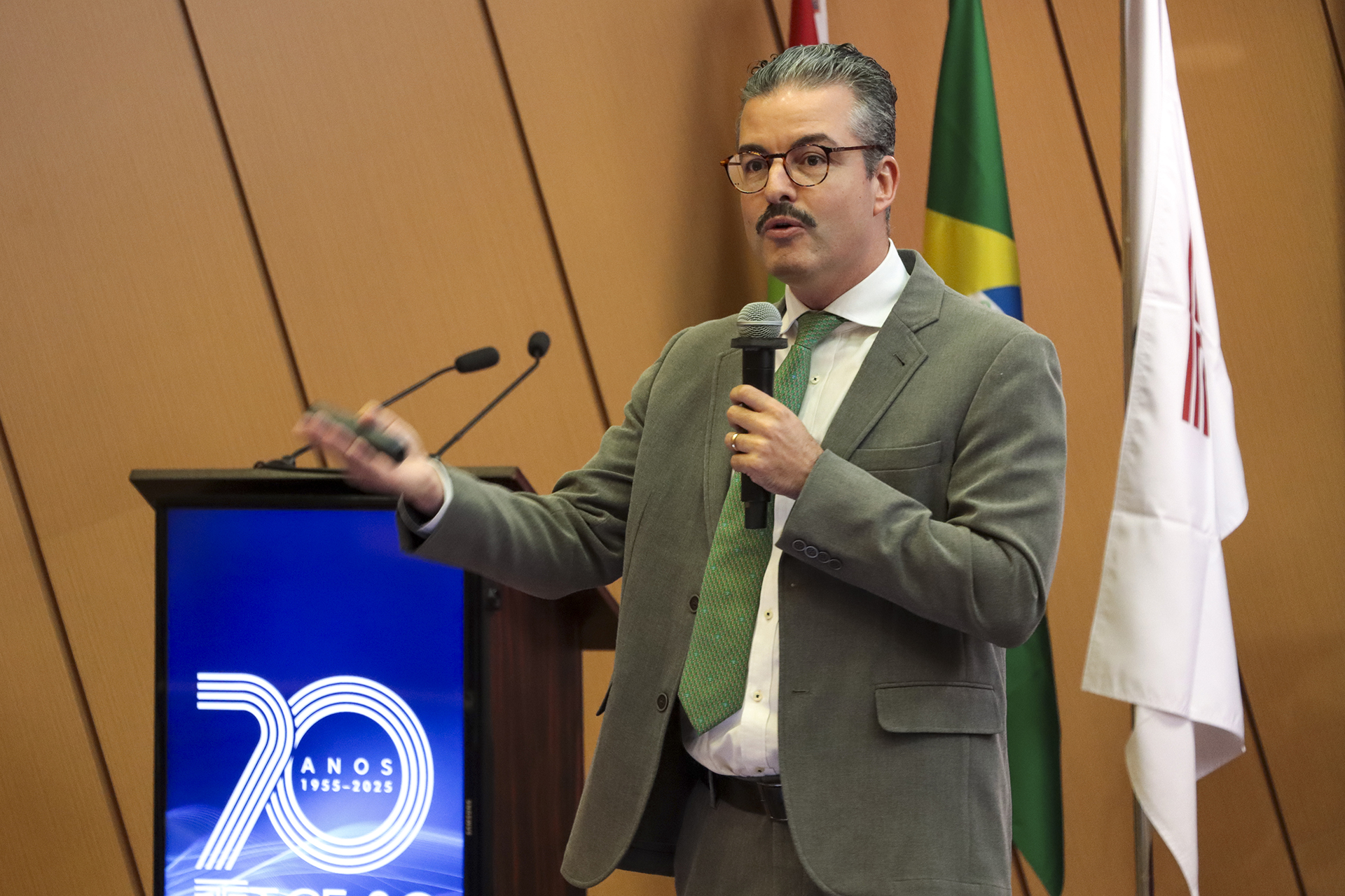 Foto horizontal que mostra o conselheiro Felipe Galvão Puccioni, do TCMRio, durante sua fala no púlpito do auditório do TCE/SC, no evento de treinamento, utilizando microfone e gesticulando enquanto se dirige ao público, em ambiente institucional decorado com bandeiras e painel comemorativo dos 70 anos do Tribunal. Ele é um homem branco, de cabelos e bigode grisalhos.