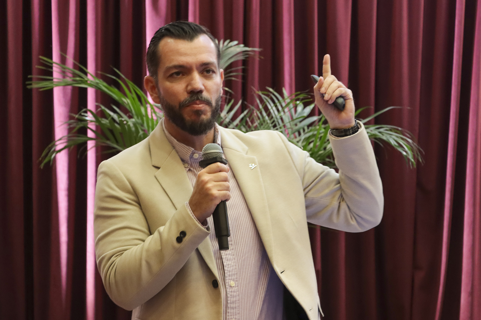 Foto horizontal que mostra o servidor do TCMRio Fabio Tessinari durante apresentação no palco do auditório do TCE/SC, no evento de treinamento, utilizando microfone e gesticulando enquanto se dirige ao público, em ambiente institucional com cortinas vermelhas e plantas decorativas ao fundo. Ele é um homem branco, de cabelos e barba escuros.