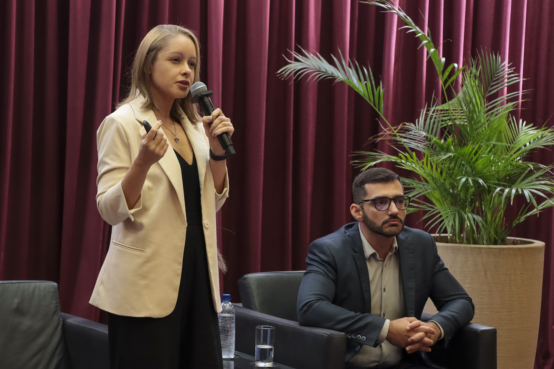 Foto horizontal que mostra a servidora do TCMRio Ana Carolina de Souza Inez durante apresentação no palco do auditório do TCE/SC, no evento de treinamento, utilizando microfone e gesticulando enquanto se dirige ao público. Ela é uma mulher branca, de cabelos claros. Ao lado, o seu colega Mario está sentado, acompanhando a exposição, em ambiente institucional com cortinas vermelhas e plantas decorativas ao fundo.