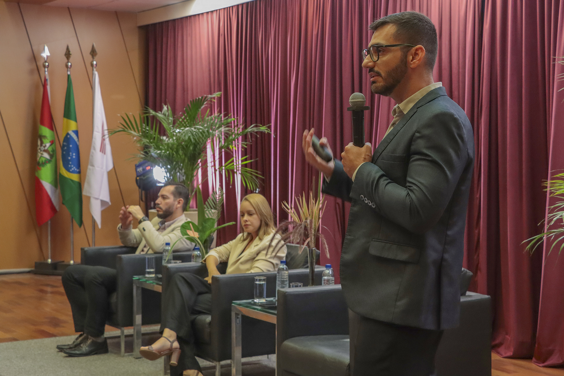 Foto horizontal de perfil do servidor do TCMRio Mario David dos Santos durante apresentação no palco do auditório do TCE/SC, no evento de treinamento, utilizando microfone e gesticulando enquanto se dirige ao público; ao fundo, seus colegas Fabio Tessinari e Ana Carolina de Souza Inez estão sentados em poltronas, acompanhando a atividade, em ambiente institucional com bandeiras, plantas decorativas e cortinas vermelhas.