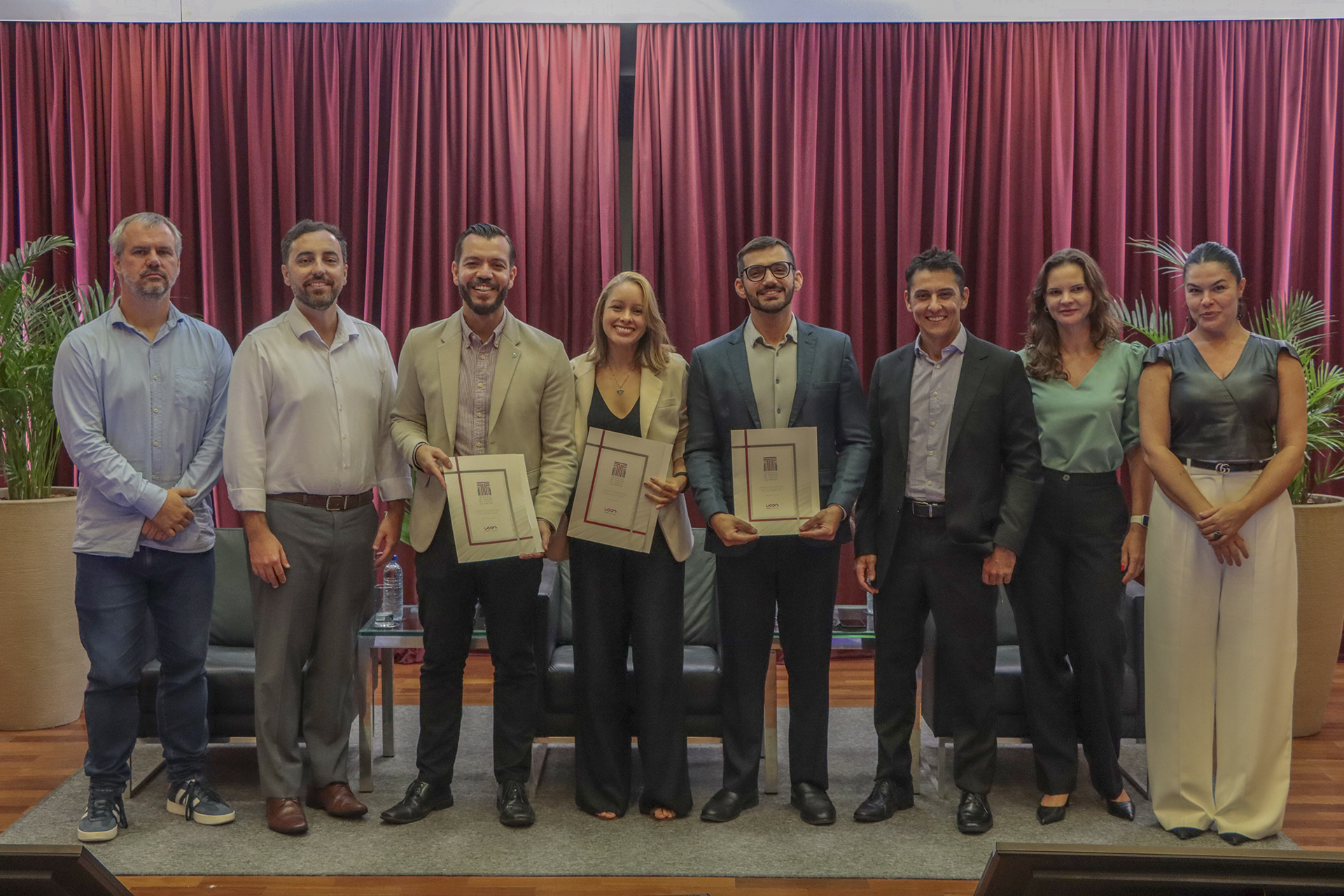 Foto horizontal que mostra autoridades e participantes lado a lado no palco do auditório do TCE/SC, durante evento de treinamento, com destaque para os servidores do TCMRio Fabio Tessinari, Mario David dos Santos Bisneto e Ana Carolina de Souza Inez, ao centro, segurando certificados da capacitação; ao lado, estão integrantes do TCE/SC, incluindo a diretora Monique Portella, em ambiente institucional com poltronas e cortinas vermelhas ao fundo.