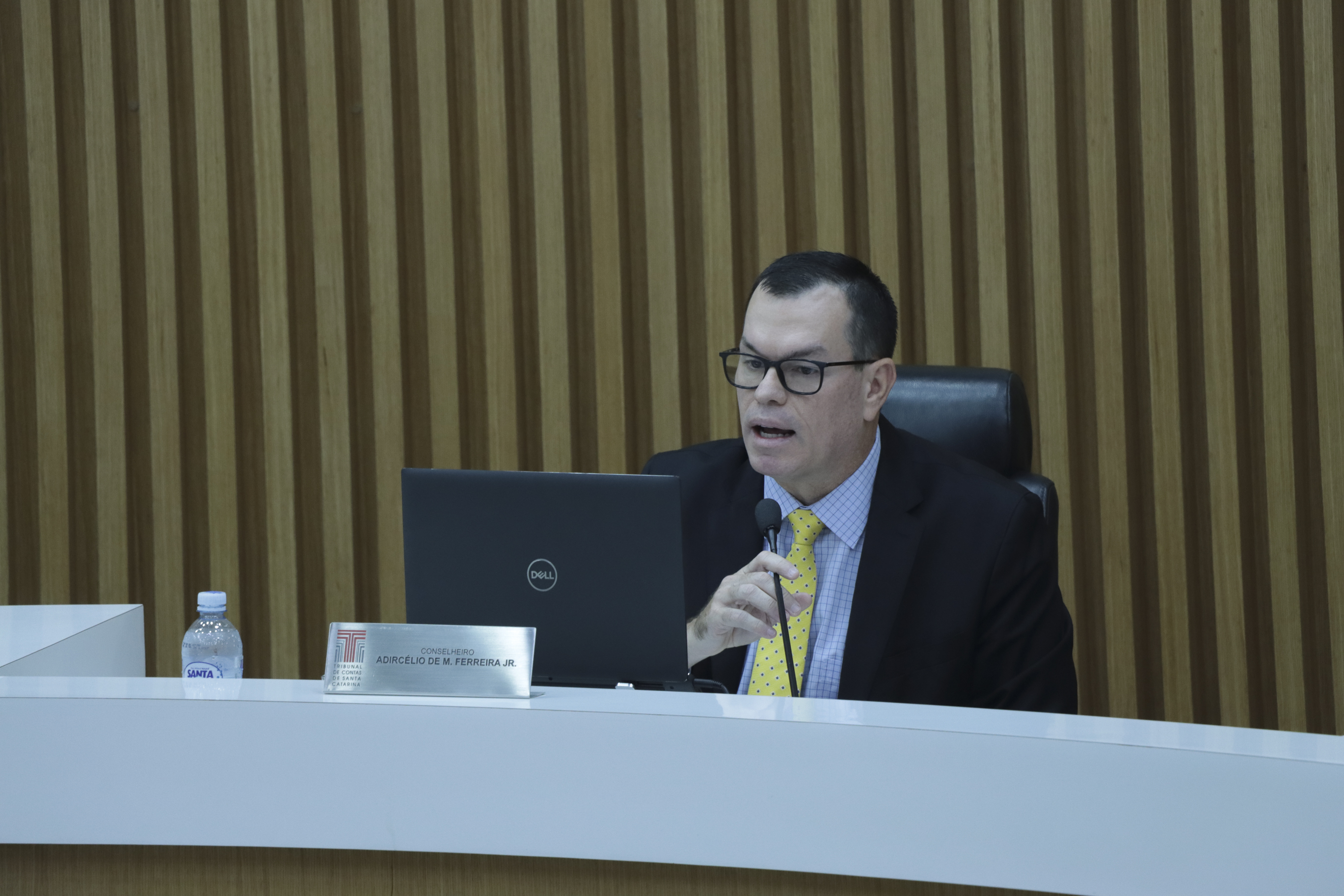 Foto horizontal do conselheiro Adircélio de Moraes Ferreira Júnior durante sessão plenária no TCE/SC, sentado à bancada, utilizando microfone e falando enquanto olha para a tela de um notebook. Ele é um homem branco, de cabelos pretos. Veste terno e gravata, usa óculos de grau e está em ambiente institucional com painel de madeira ao fundo.