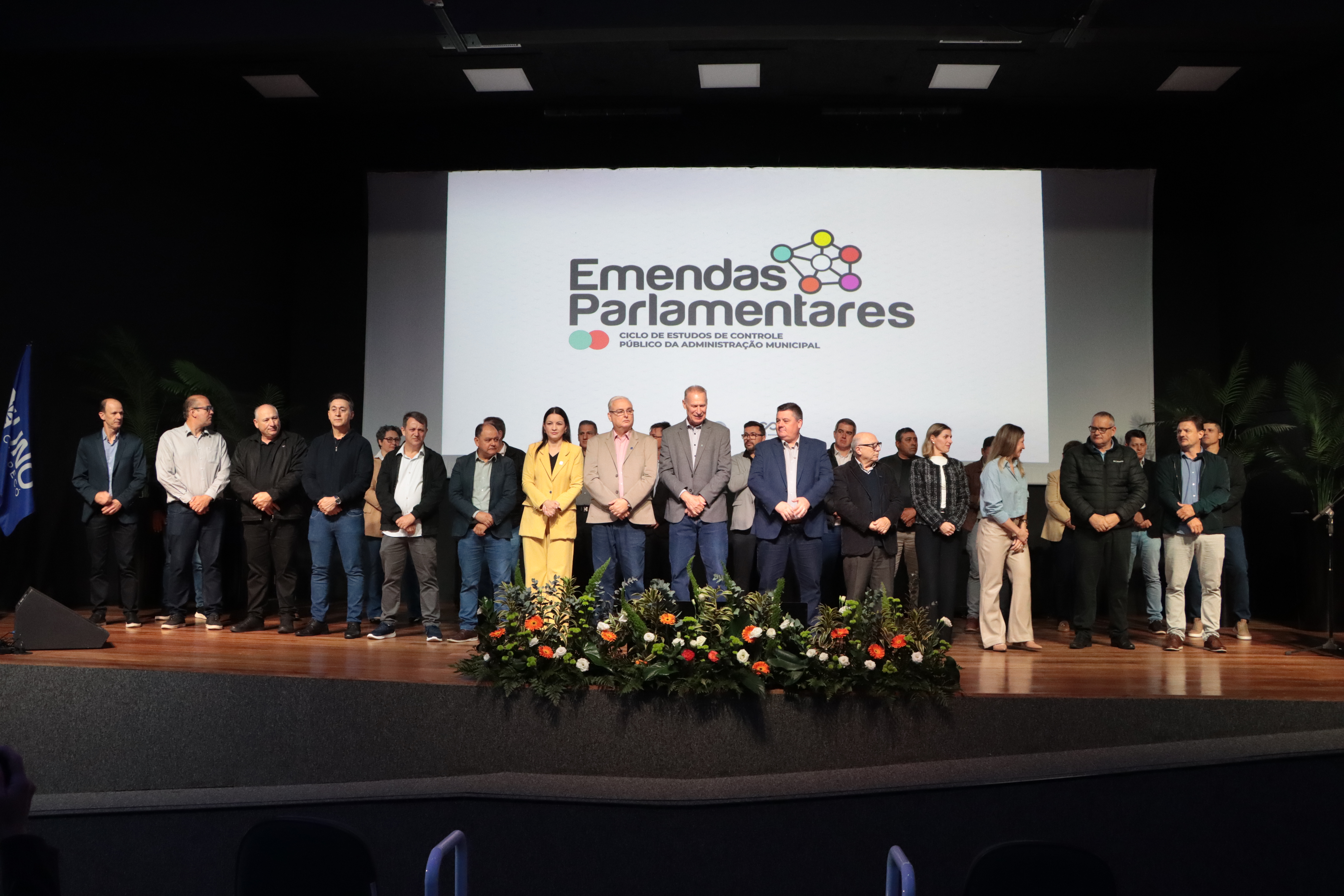 Foto horizontal da comitiva do TCE/SC posicionada no palco do auditório. Há várias pessoas, lado a lado. À frente, o presidente Herneus e diretores do TCE/SC. Ao fundo, no telão, logo do Ciclo de Estudos sobre Emendas Parlamentares.