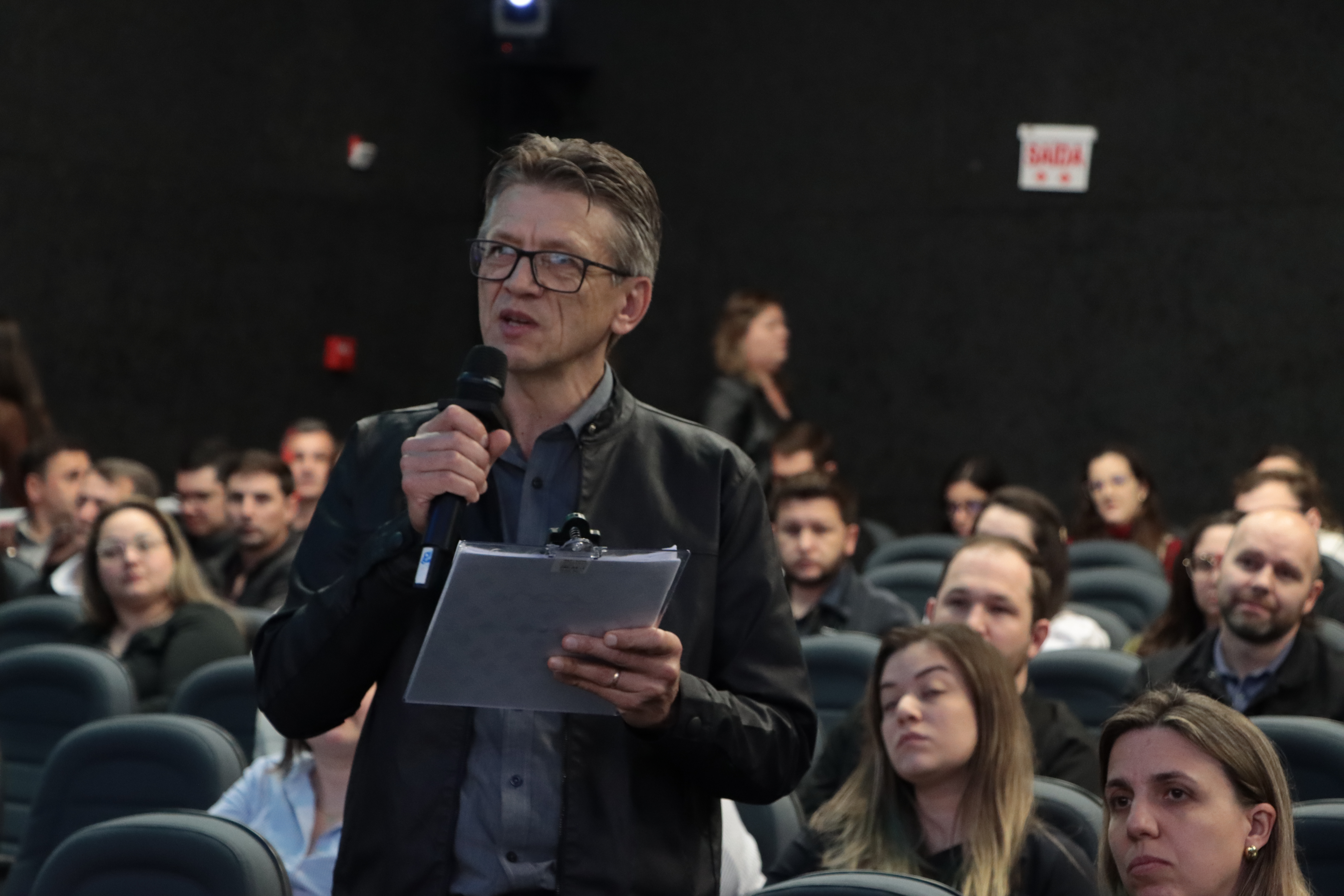 Foto horizontal de um dos participantes do evento fazendo uma pergunta aos palestrantes. Ele está em pé em meio ao restante da plateia, segurando um papel e falando ao microfone.
