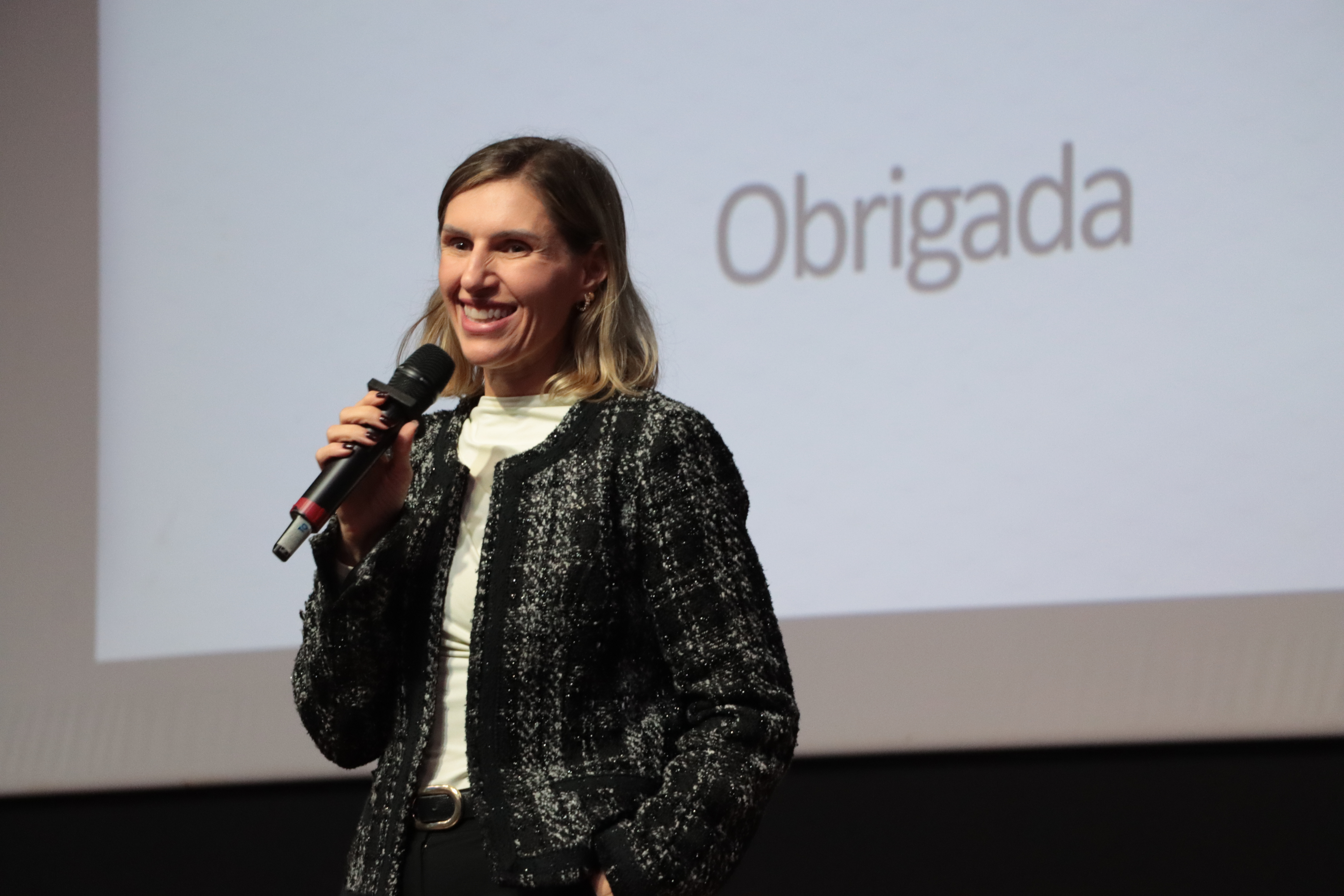 Foto horizontal da diretora de Gestão de Contas (DGE), Claudia Vieira da Silva, falando ao microfone durante o evento. É uma mulher branca, de cabelos claros. Está em pé e sorri. Ao fundo, projeção de slides.