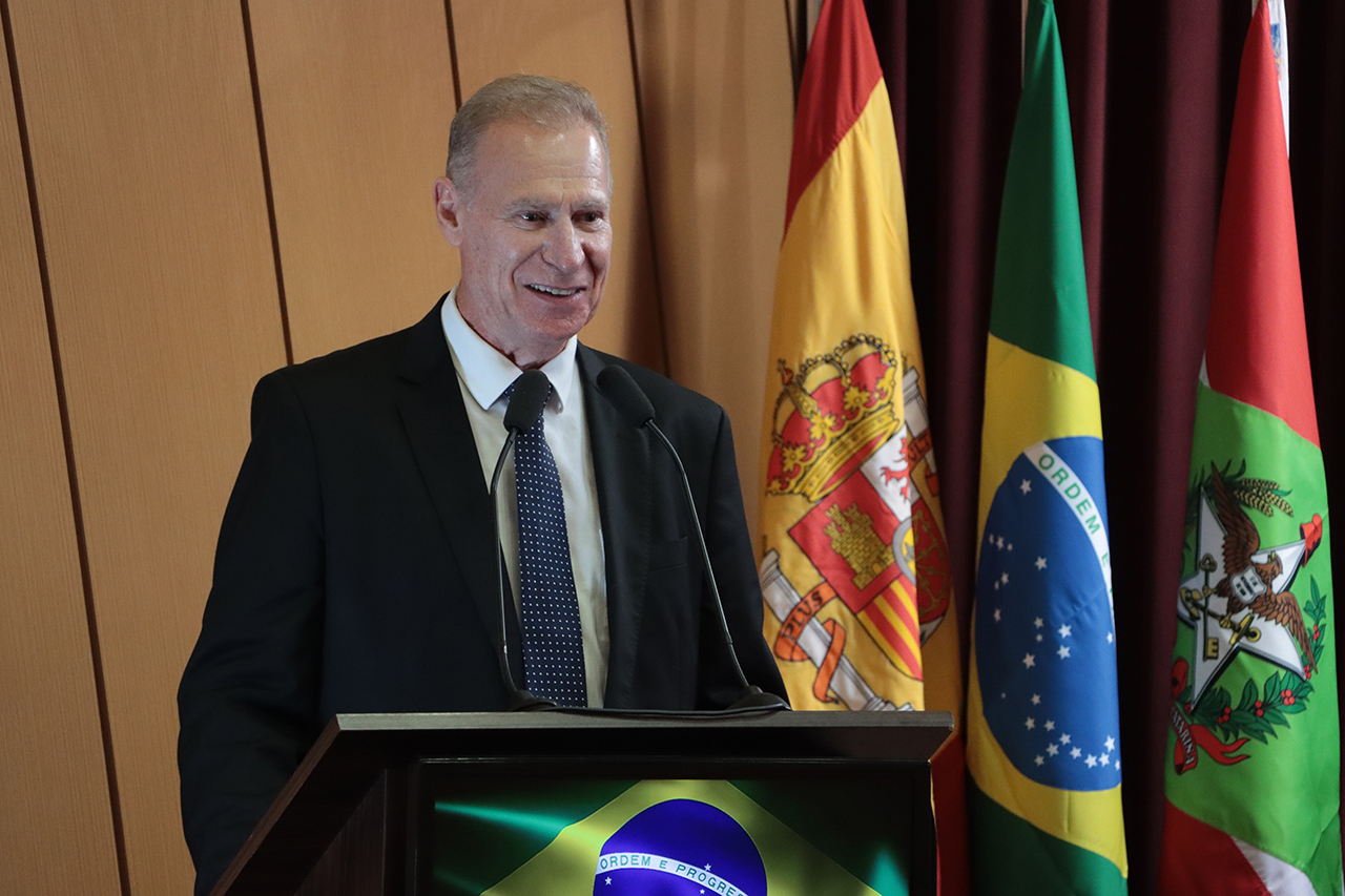 Foto do presidente do TCE/SC, conselheiro Herneus De Nadal, discursando no púlpito do auditório durante o evento. Ele é um homem branco, de cabelos claros. Veste terno e gravata. Ao lado, no palco, várias autoridades.