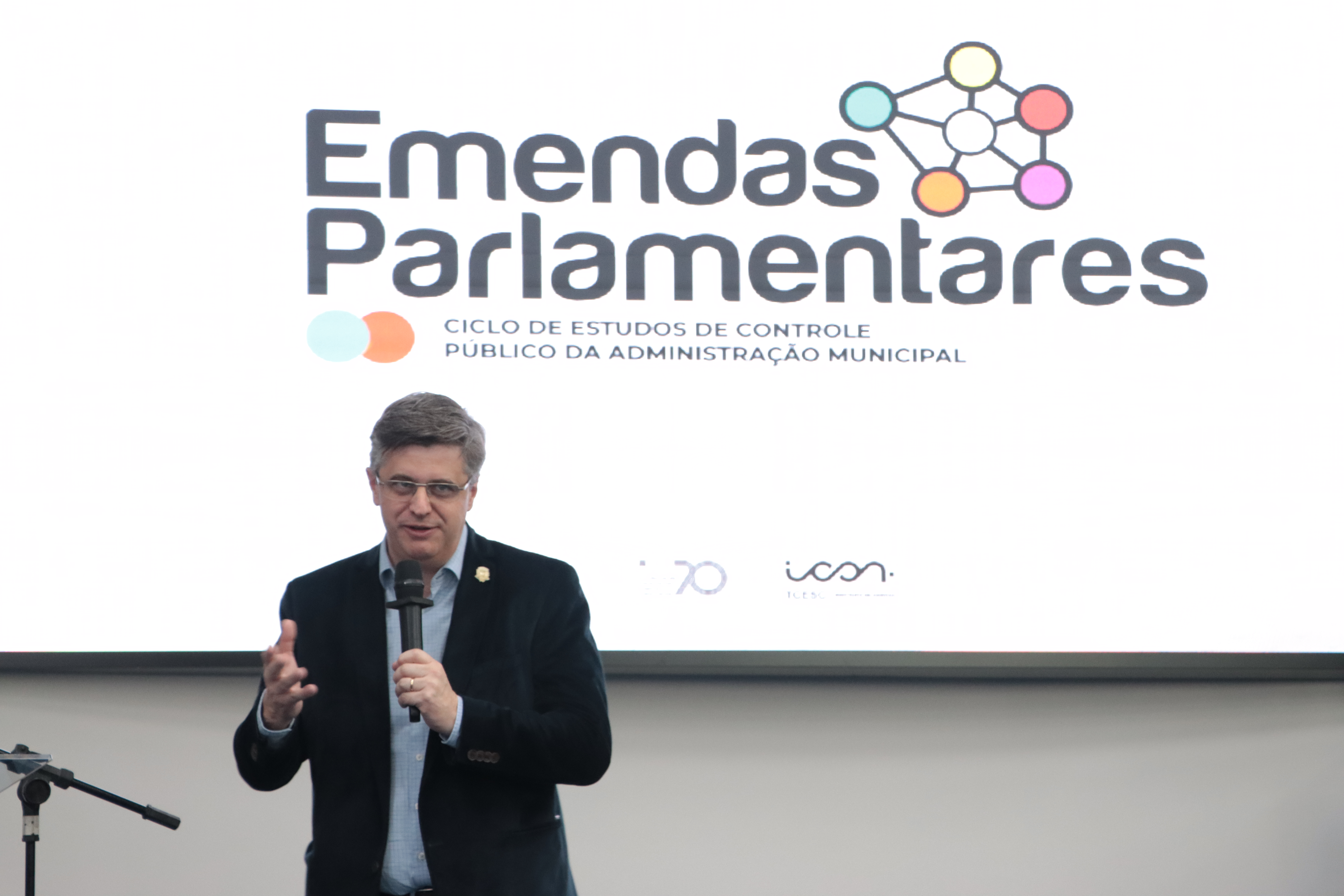 Foto horizontal do prefeito de Caçador, Alencar Mendes, falando ao microfone no palco do auditório. Ao fundo, telão exibe logo do Ciclo sobre emendas parlamentares.