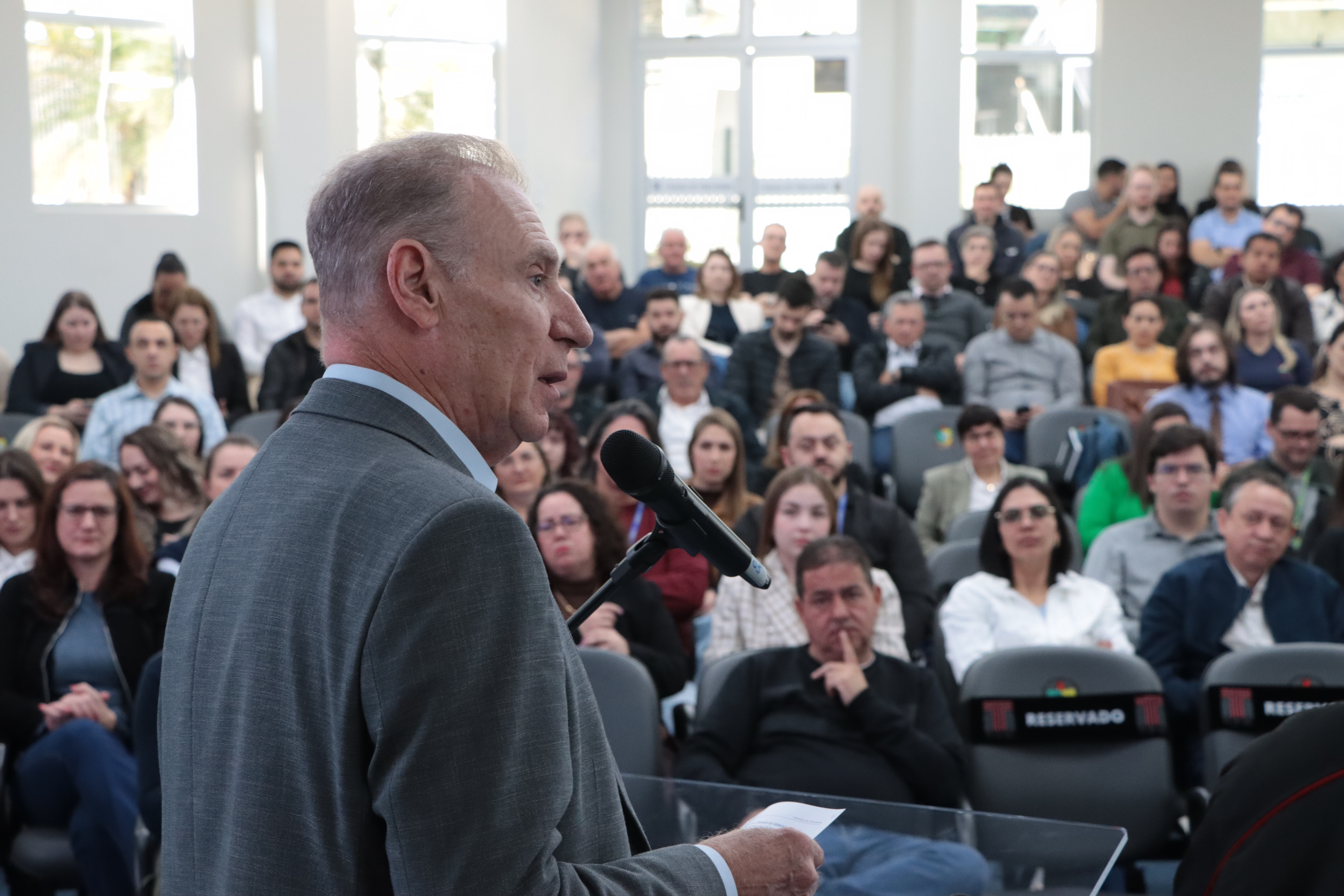 Foto horizontal do presidente Herneus De Nadal discursando no púlpito do auditório. Ele é um homem branco, de cabelos claros. Veste paletó cinza. À sua frente, várias pessoas assistem atentamente.