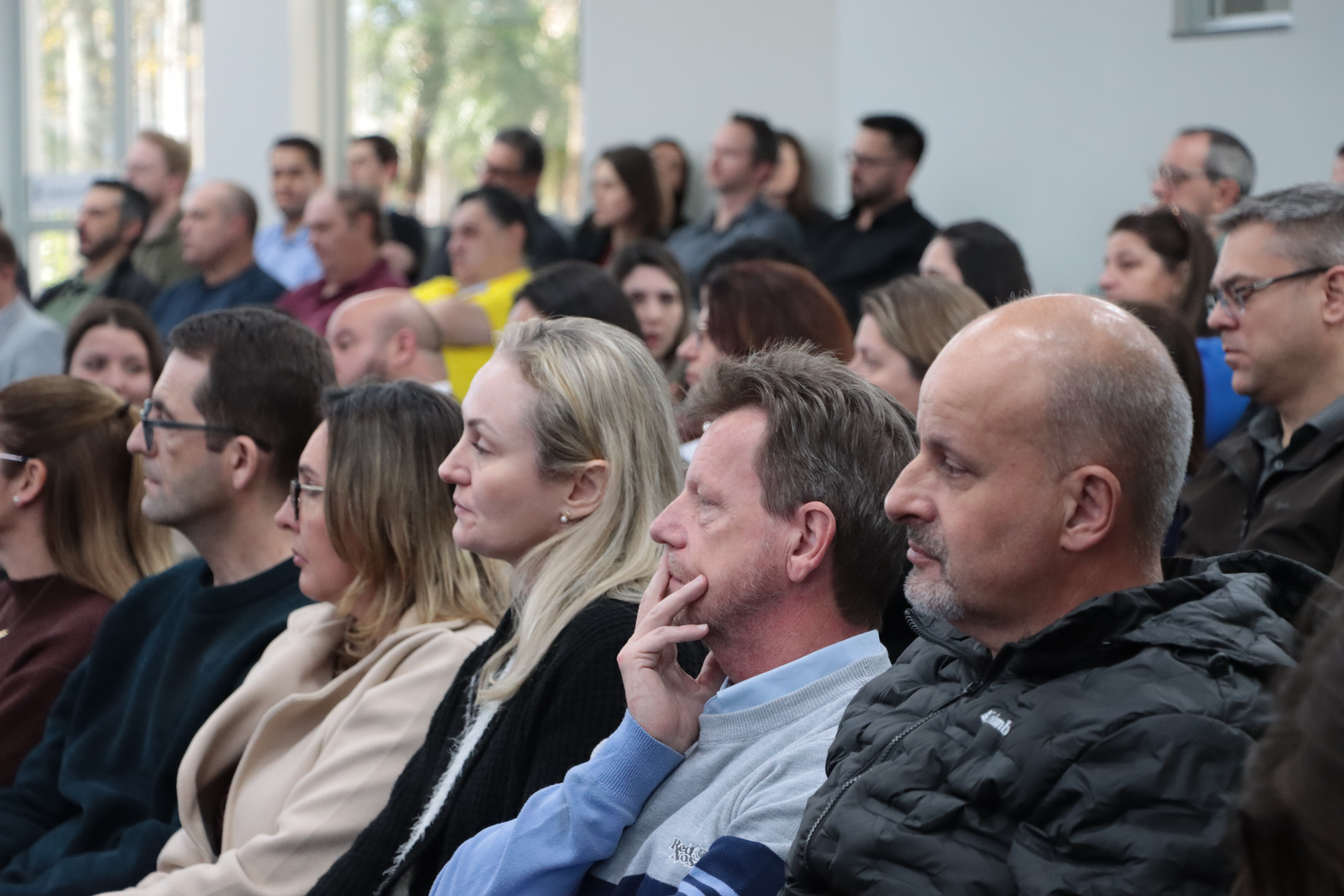 Foto horizontal do público participante sentado em poltronas do auditório. Eles escutam atentamente.