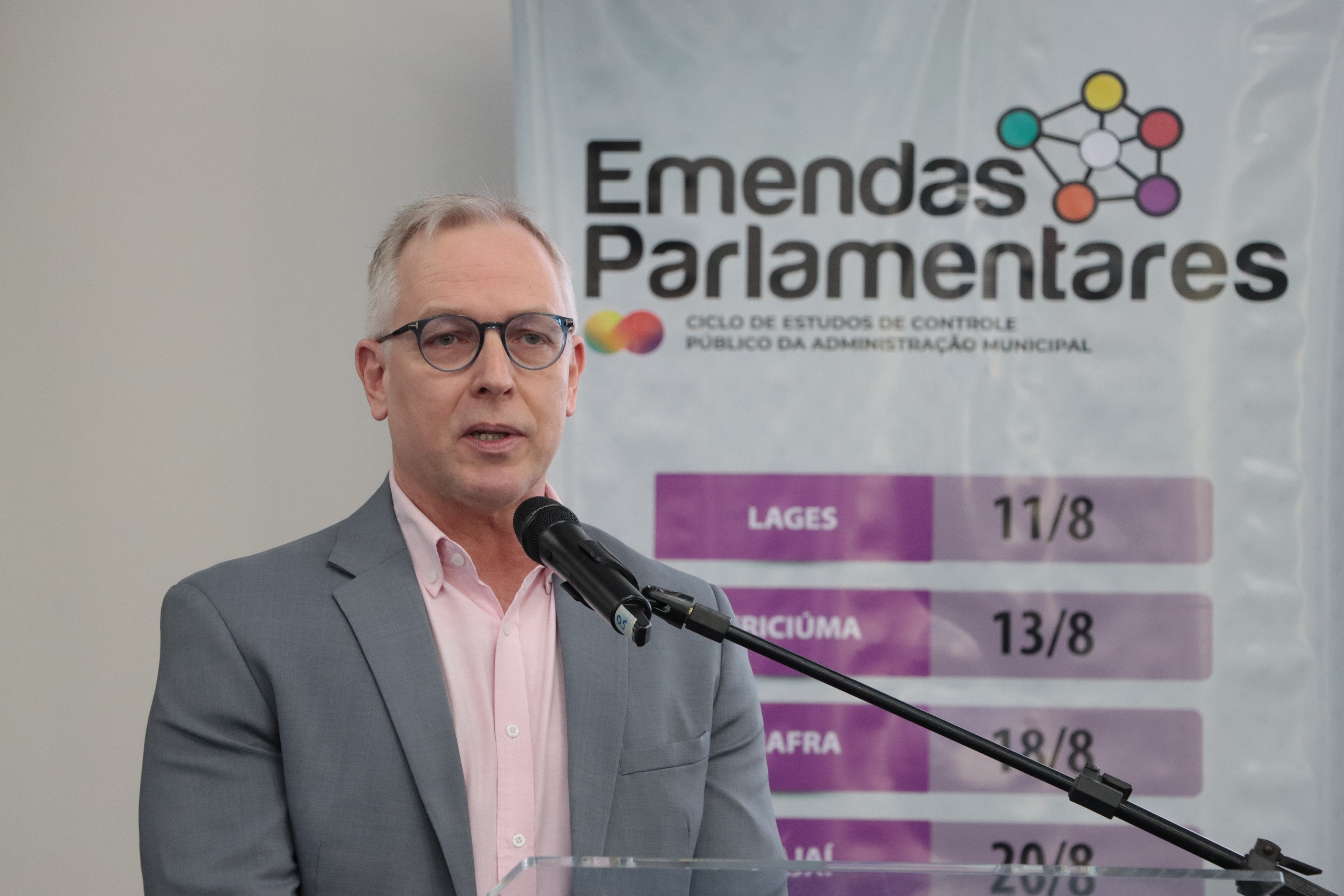 Foto horizontal do diretor Sidney Tavares Júnior falando ao microfone. Ele é um homem branco, de cabelos grisalhos. Veste paletó cinza.