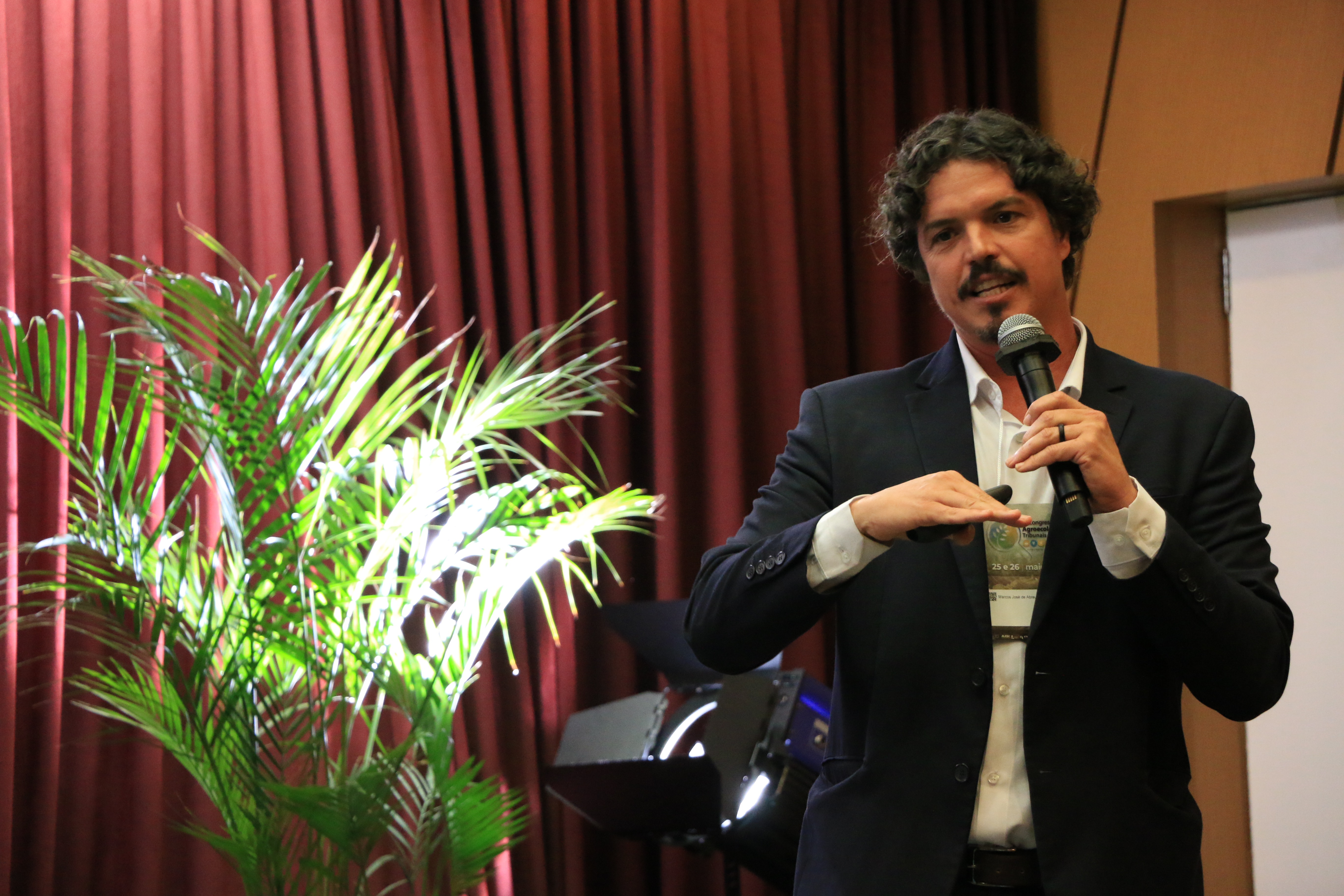 Imagem horizontal do deputado estadual Marquito. Ele é um homem branco, de cabelos castanhos e cacheados. Está em pé, segurando um microfone e um controle de apresentação, usando camisa paletó preto e crachá do evento. Ao fundo, cortinas bordôs e uma planta ornamental em vaso compõem o cenário do auditório do TCE/SC.