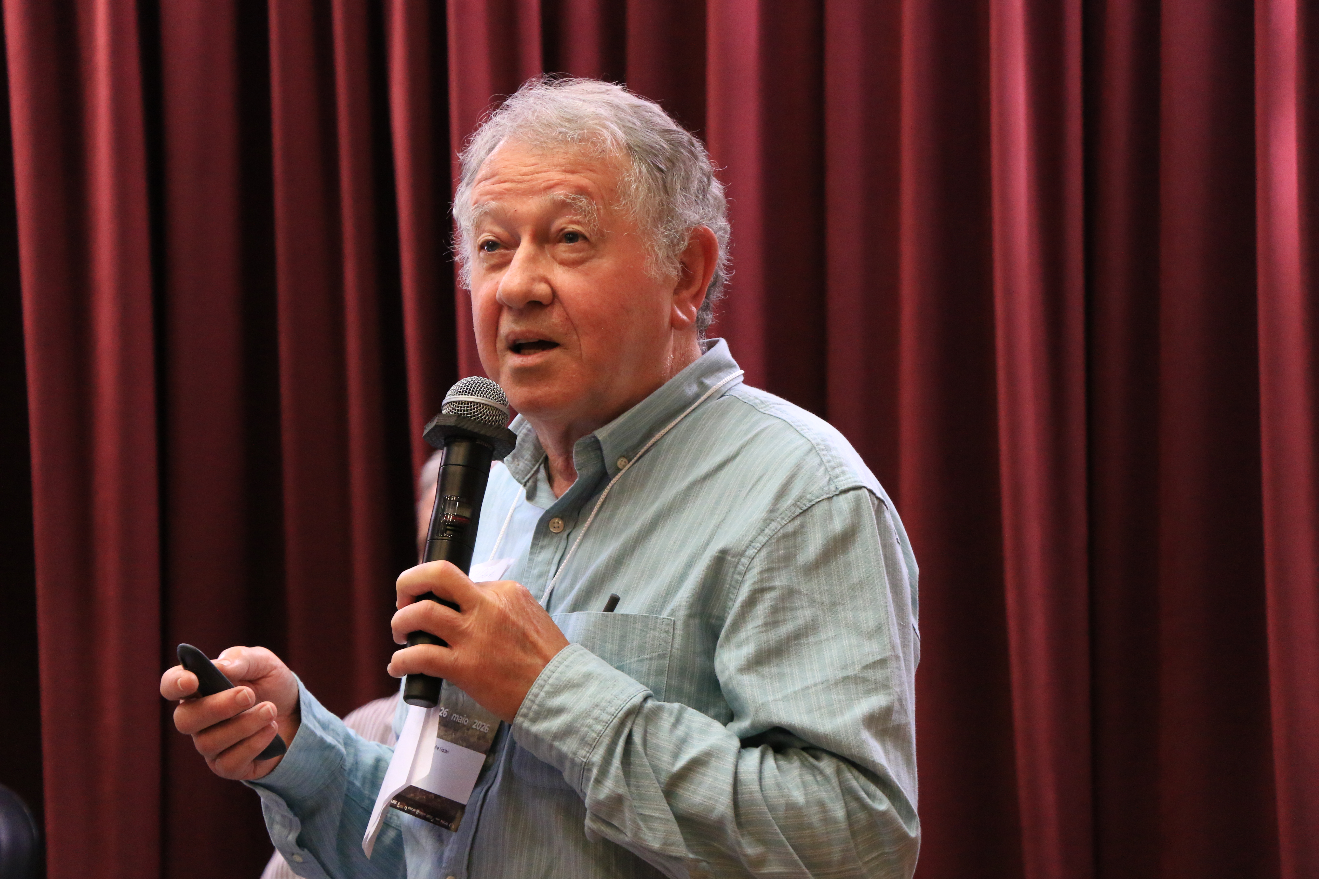 Foto horizontal do professor Rubens Nodari falando ao microfone durante o evento. É um homem branco, de cabelos grisalhos. Veste camisa social clara e segura o controle de apresentação. Ao fundo, há cortinas bordôs.