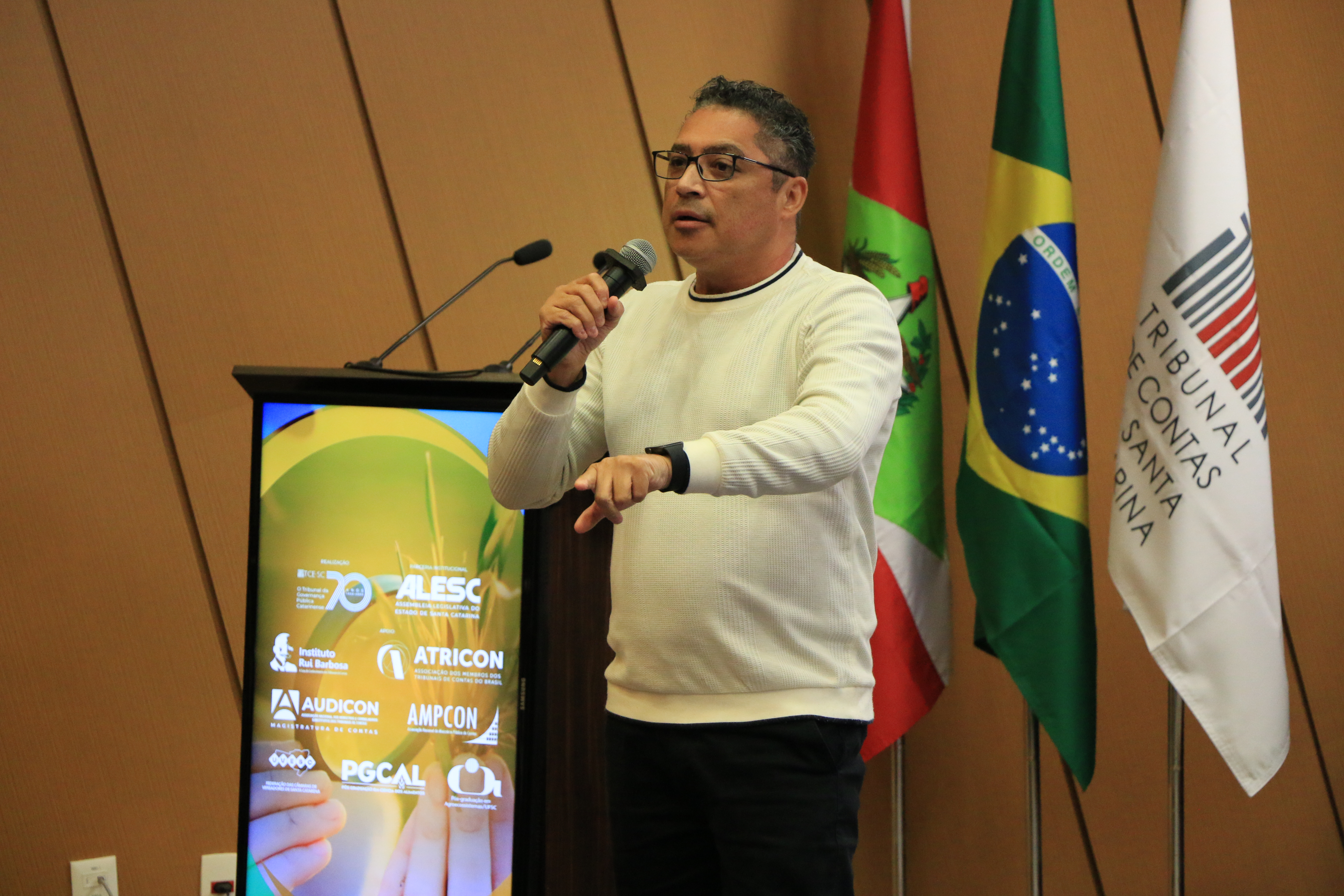 Foto horizontal do painelista Rogério Macedo falando ao microfone no palco do auditório do TCE/SC durante evento. É um homem pardo, de cabelos escuros. Veste suéter claro e está em pé, próximo ao púlpito. Ao fundo, painel amadeirado e bandeiras institucionais.