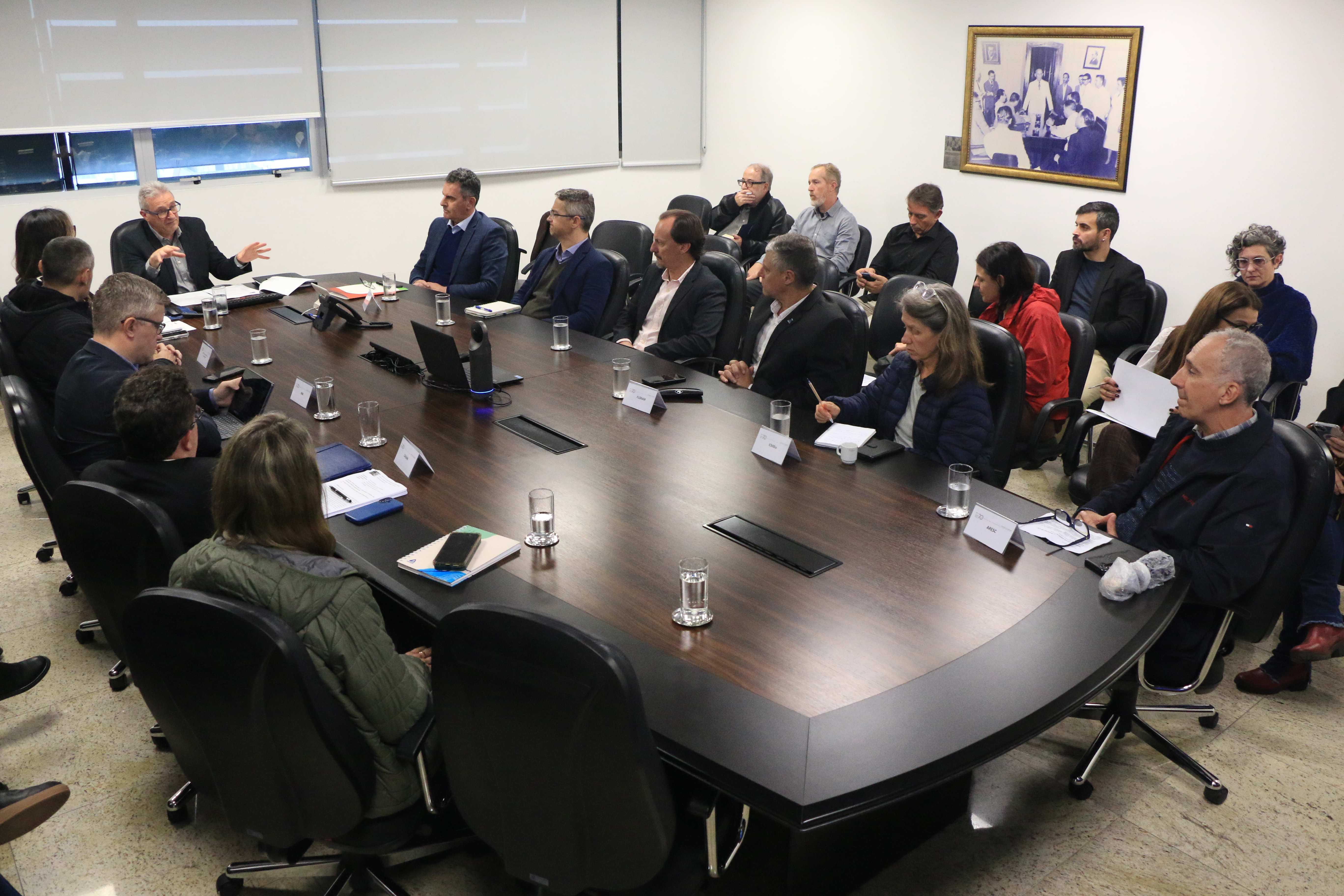 Foto horizontal ampla que mostra os participantes da mesa de consensualismo durante reunião. Há várias pessoas sentadas lado a lado diante de uma mesa. Na ponta, o coordenador, Neimar Paludo. 