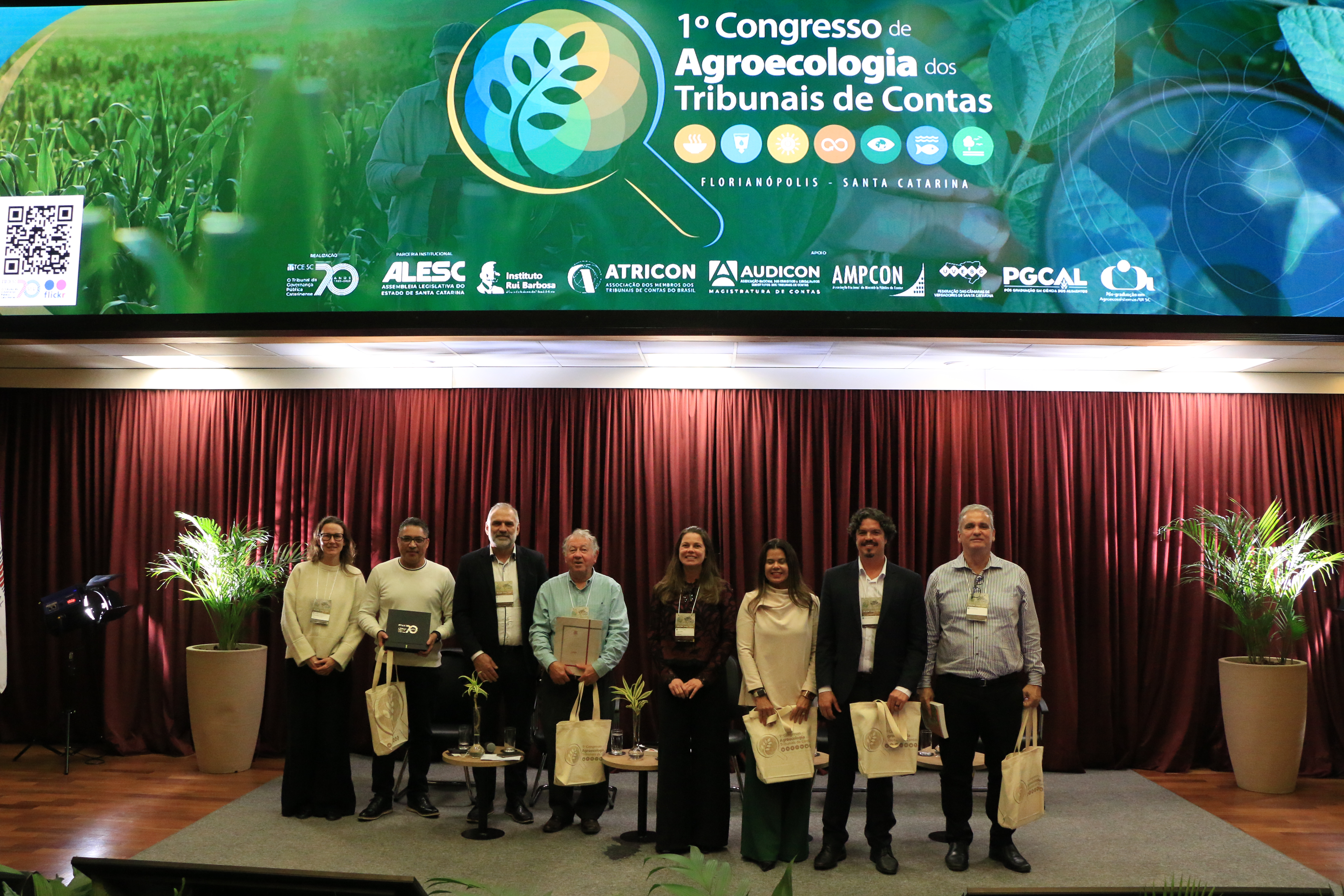 Imagem horizontal com foto posada dos participantes de um dos painéis, lado a lado, no palco do auditório do TCE/SC. Entre os presentes, a procuradora Cibelly Farias, a conselheira substituta Sabrina Iocken, o deputado estadual Marquito, e os professores Rubens Nodari e Rogério Macedo. Acima do palco, um grande painel eletrônico exibe a identidade visual do 1º Congresso de Agroecologia dos Tribunais de Contas.