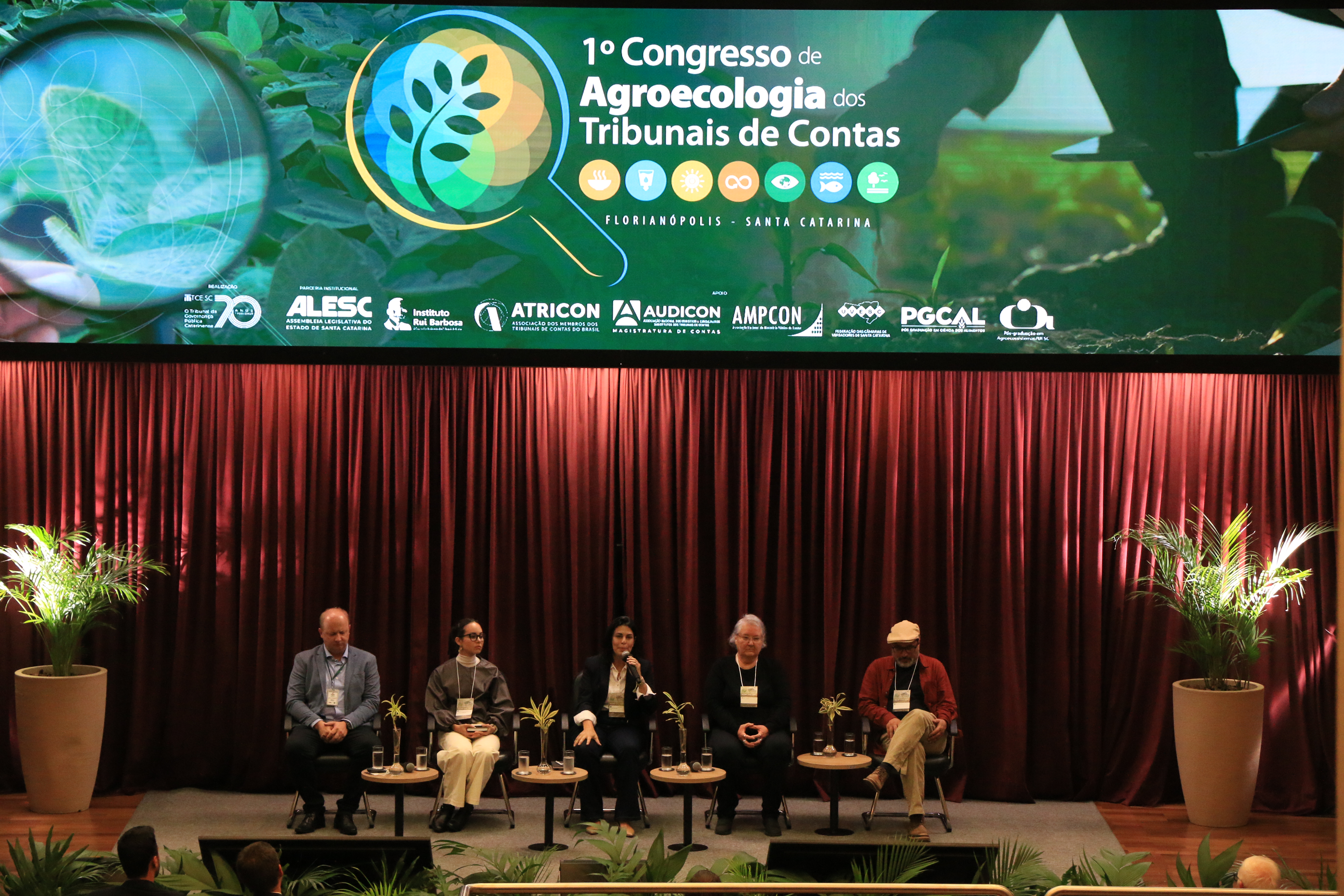 Foto horizontal dos cinco participantes do painel sentados lado a lado em poltronas no palco do auditório do TCE/SC. Entre eles, a professora Sonia Hess e a mediadora, presidente da Uvesc, Marcieli Vignatti.