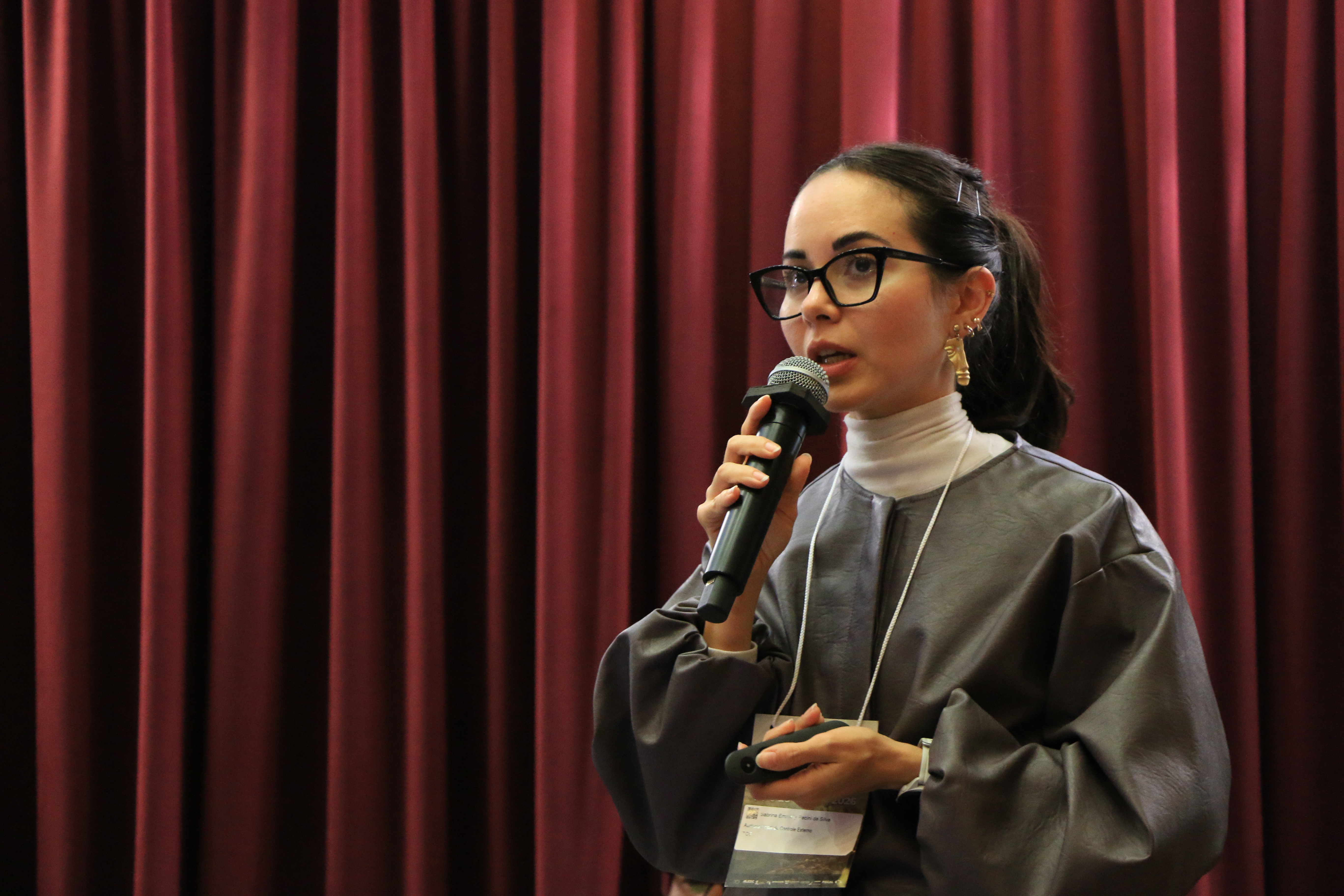 Foto horizontal da auditora do TCE/SC Sabrina Emmelly Pecini da Silva falando ao microfone. Ela é uma mulher branca, de cabelos escuros, presos. Veste roupa cinza e usa óculos de grau. Ao fundo, cortinas bordôs.